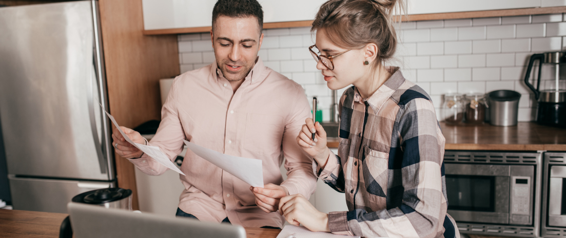 A man and a woman sit at a kitchen table reviewing paperwork in front of a laptop.