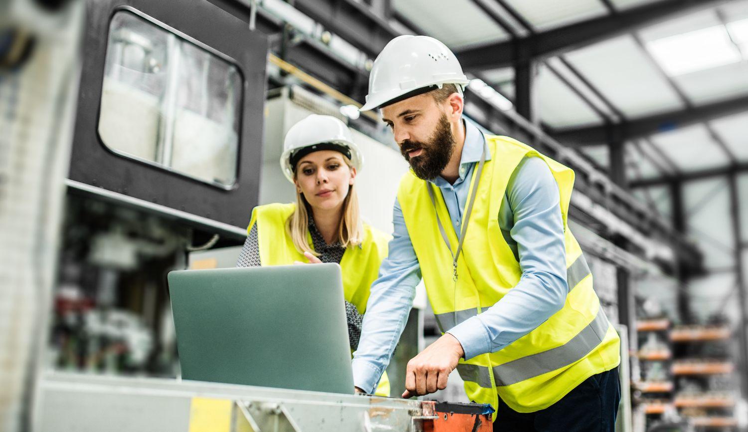 A man and a woman wearing hard hats and safety vests look at a laptop inside of a manufacturing facility.