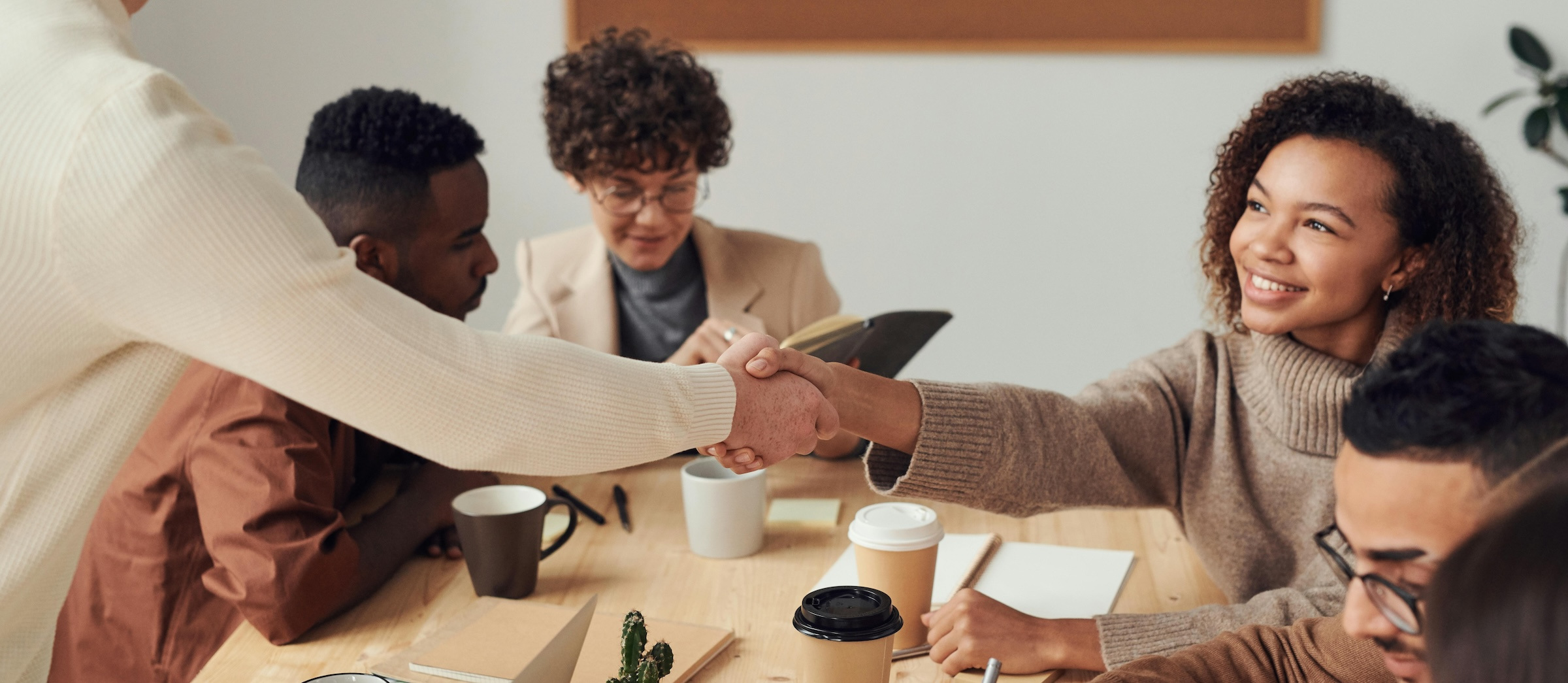 A woman shakes hands with another while seated at a conference table surrounded by other employees.