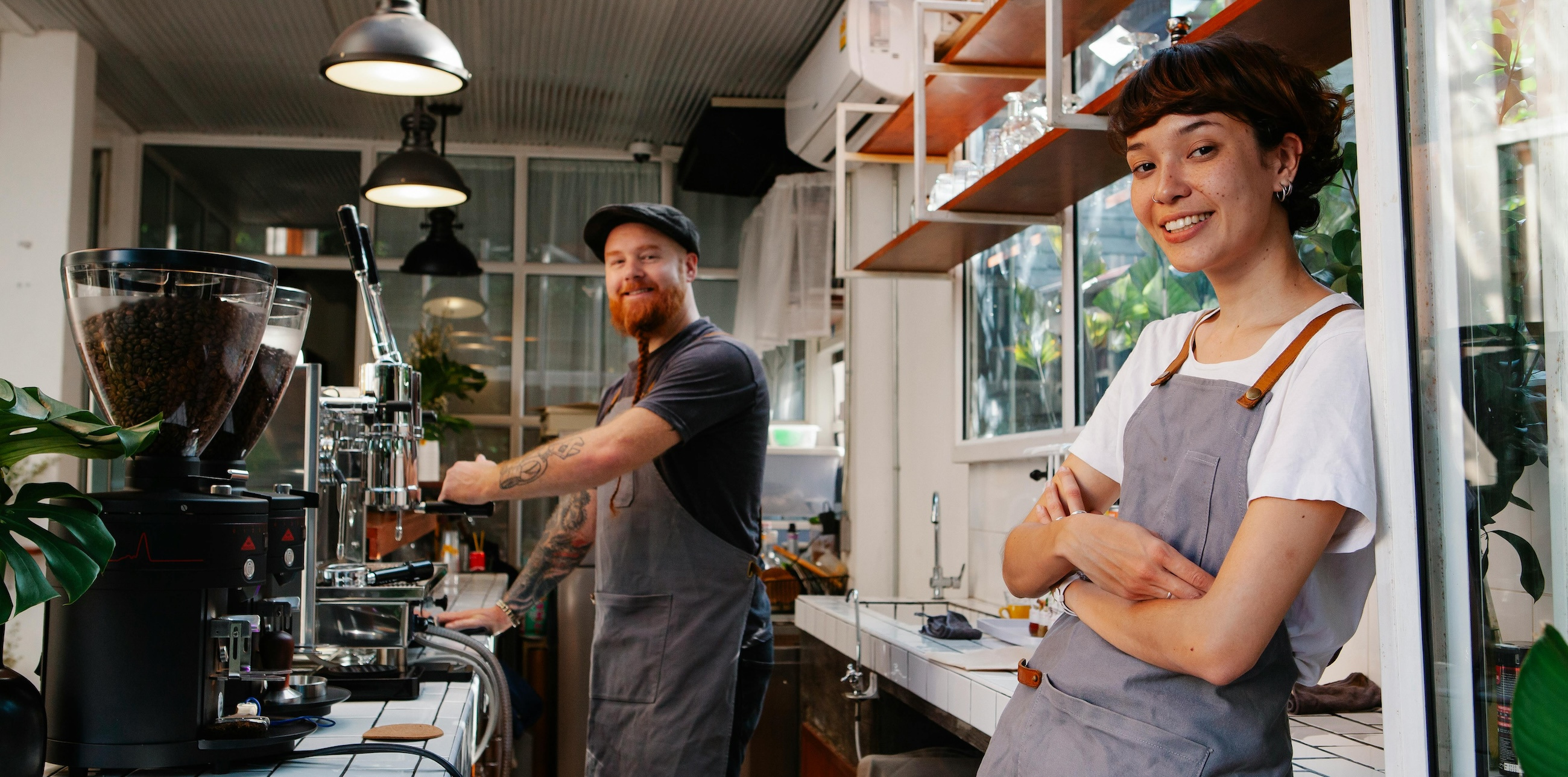 Two baristas smile at the camera behind a counter filled with coffee making equipment.