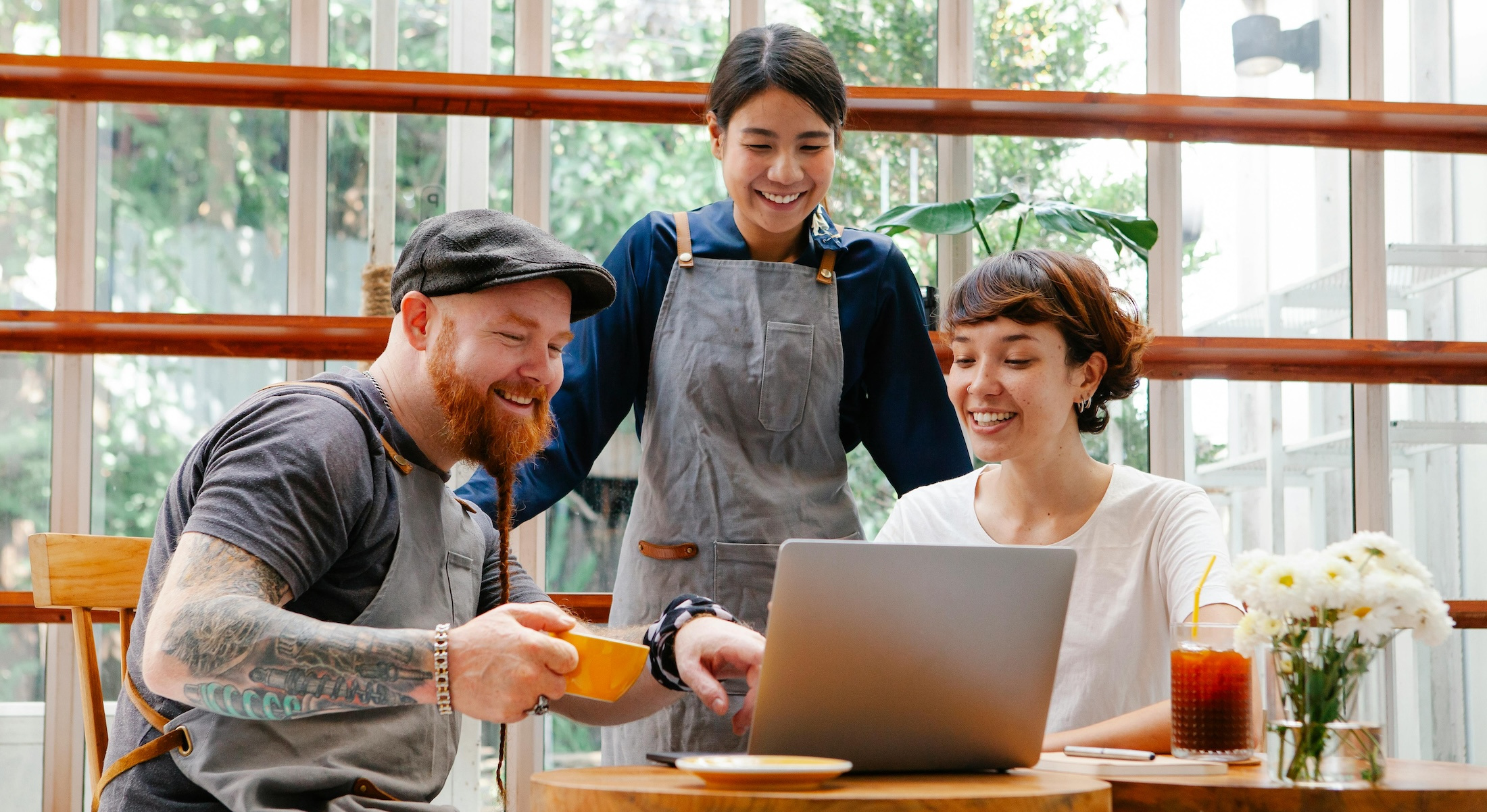 Three coffee shop employees look at a laptop screen together, smiling.