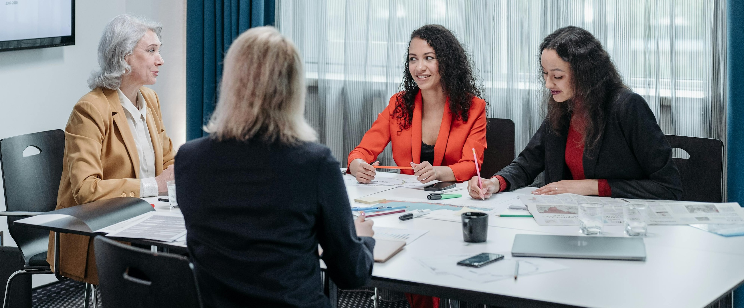 A group of 4 businesswomen sit around a conference table talking and smiling.