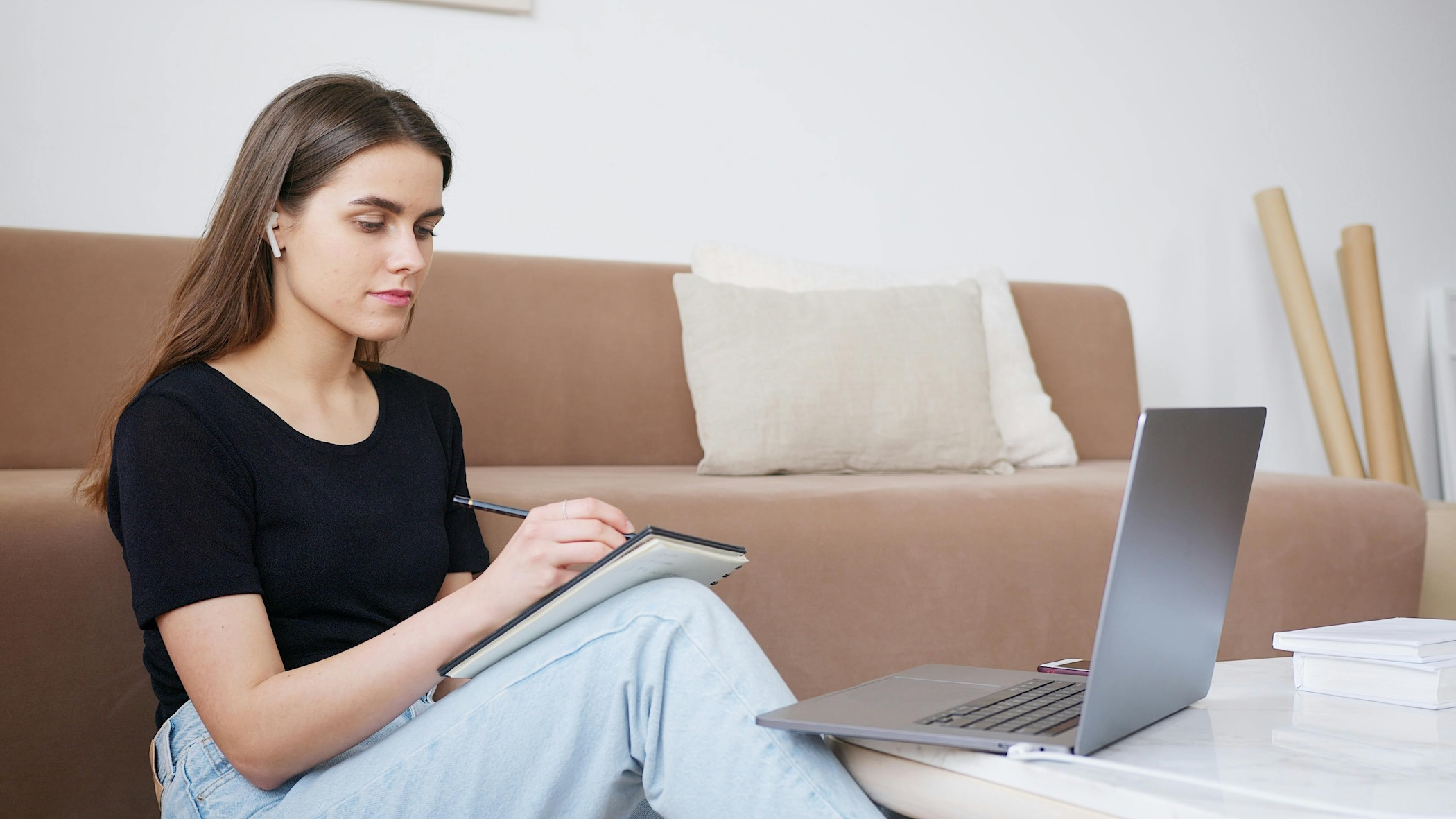 Young woman site writing in a notebook with a laptop open on the table in front of her. She wears earbuds.