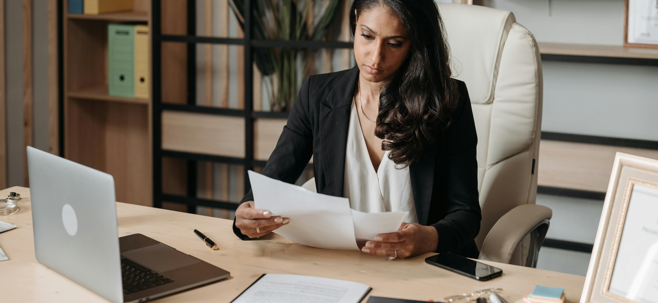 Woman in business attire sits at a desk with an open laptop and reviews paperwork.