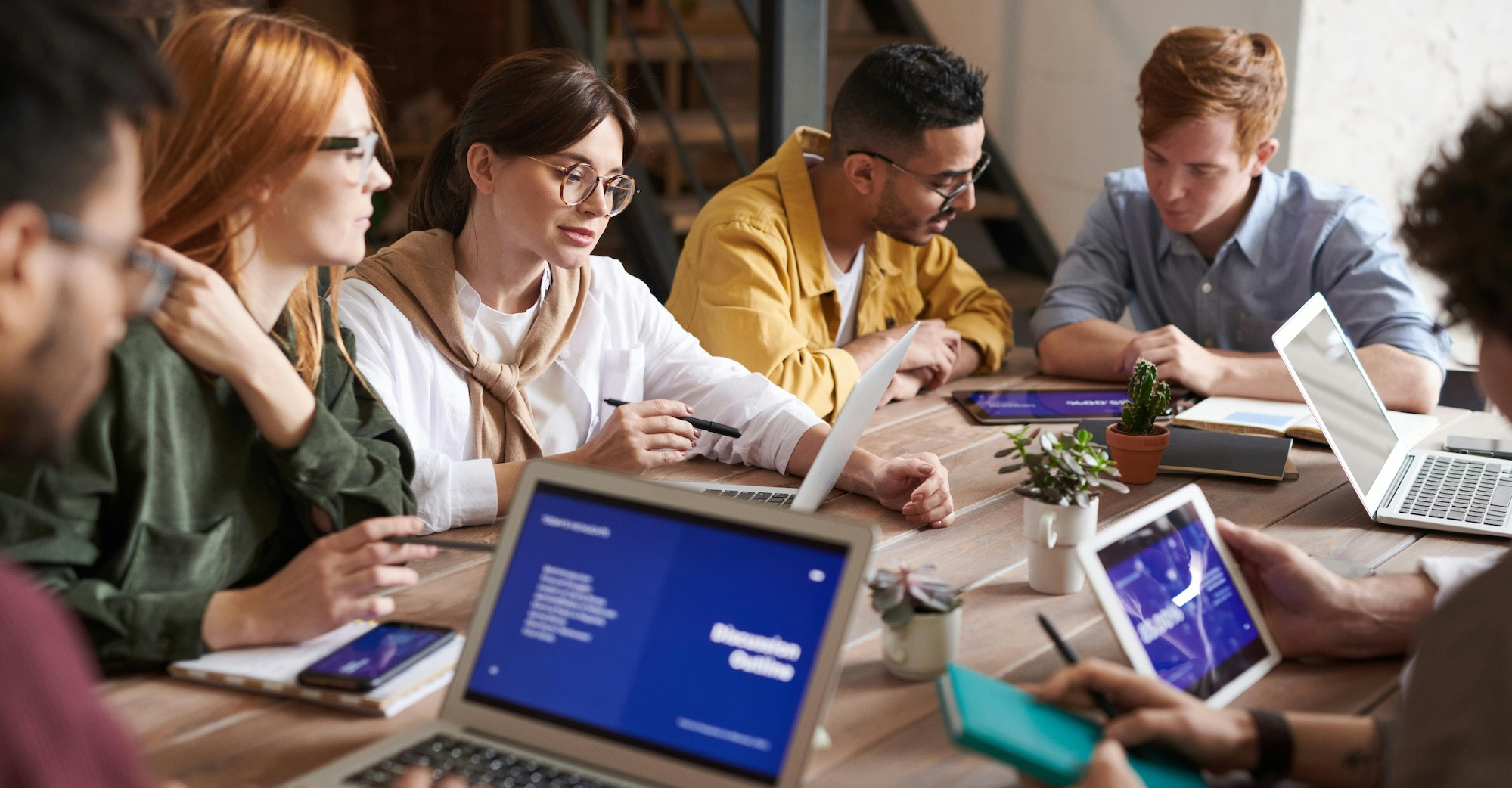 A group of employees collaborates with laptops and paperwork at a table together.