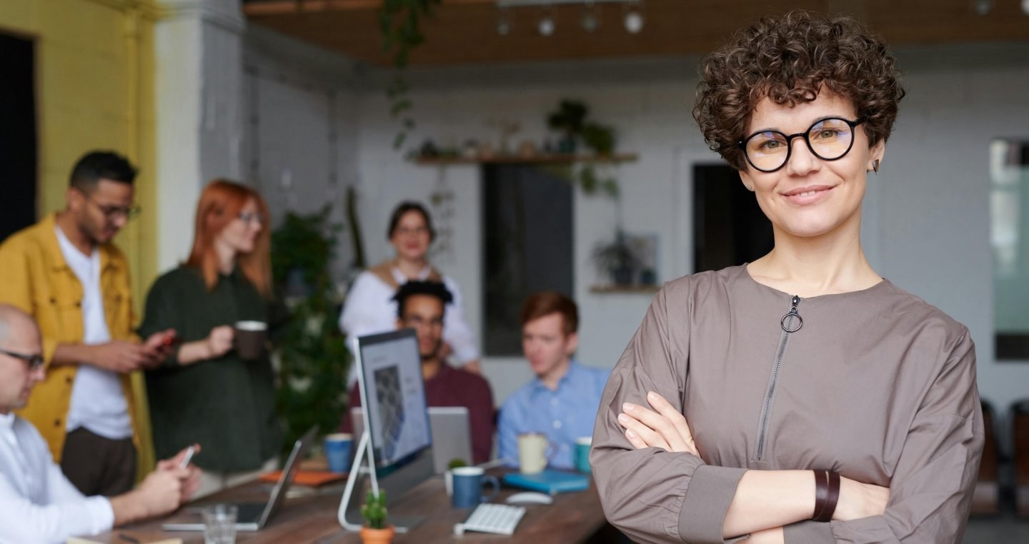A woman wearing glasses stands in the foreground crossing her arms and smiling with a group of employees working behind her.