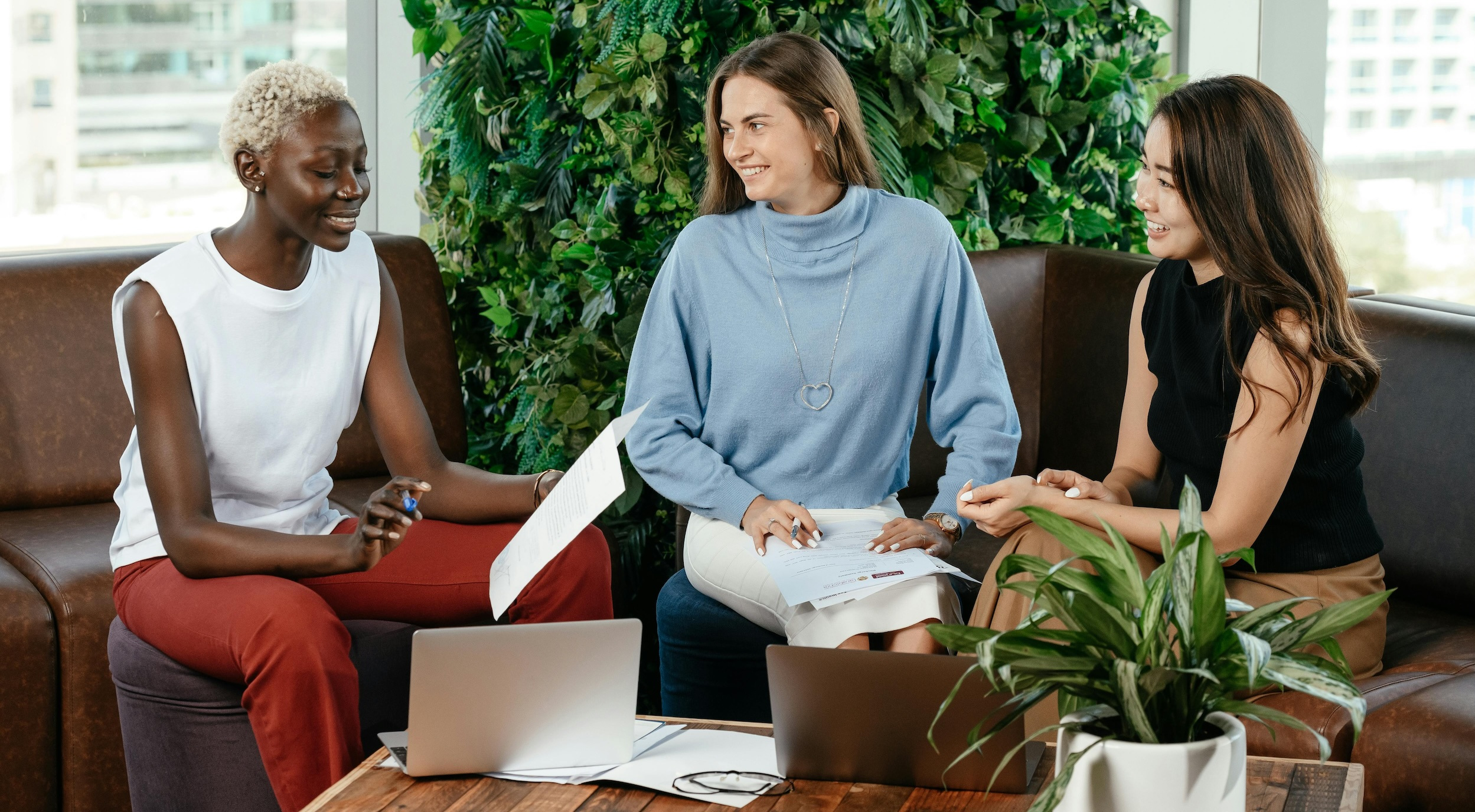 Three women sitting on couches in an office talking while reviewing papers with laptops open in front of them.