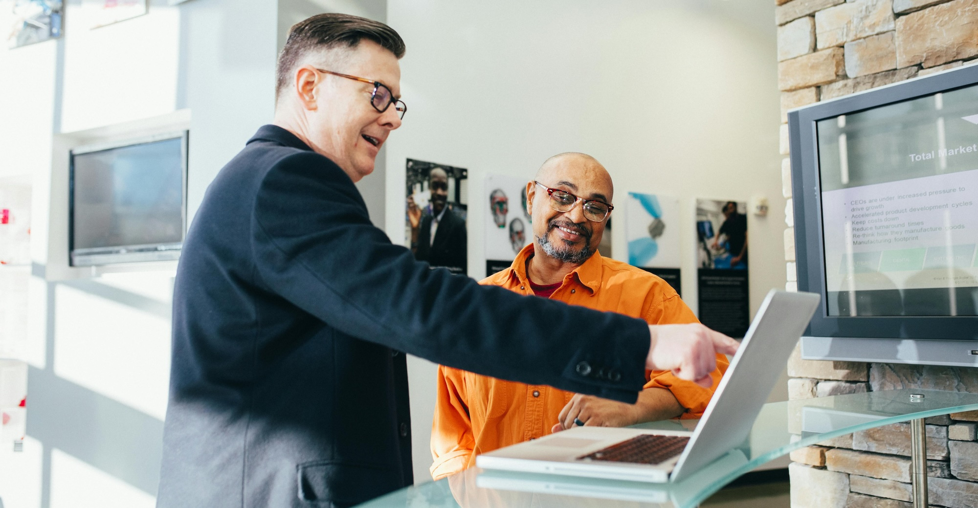 A man in a business suit points to a laptop screen and smiles, talking to another man looking at the same screen.