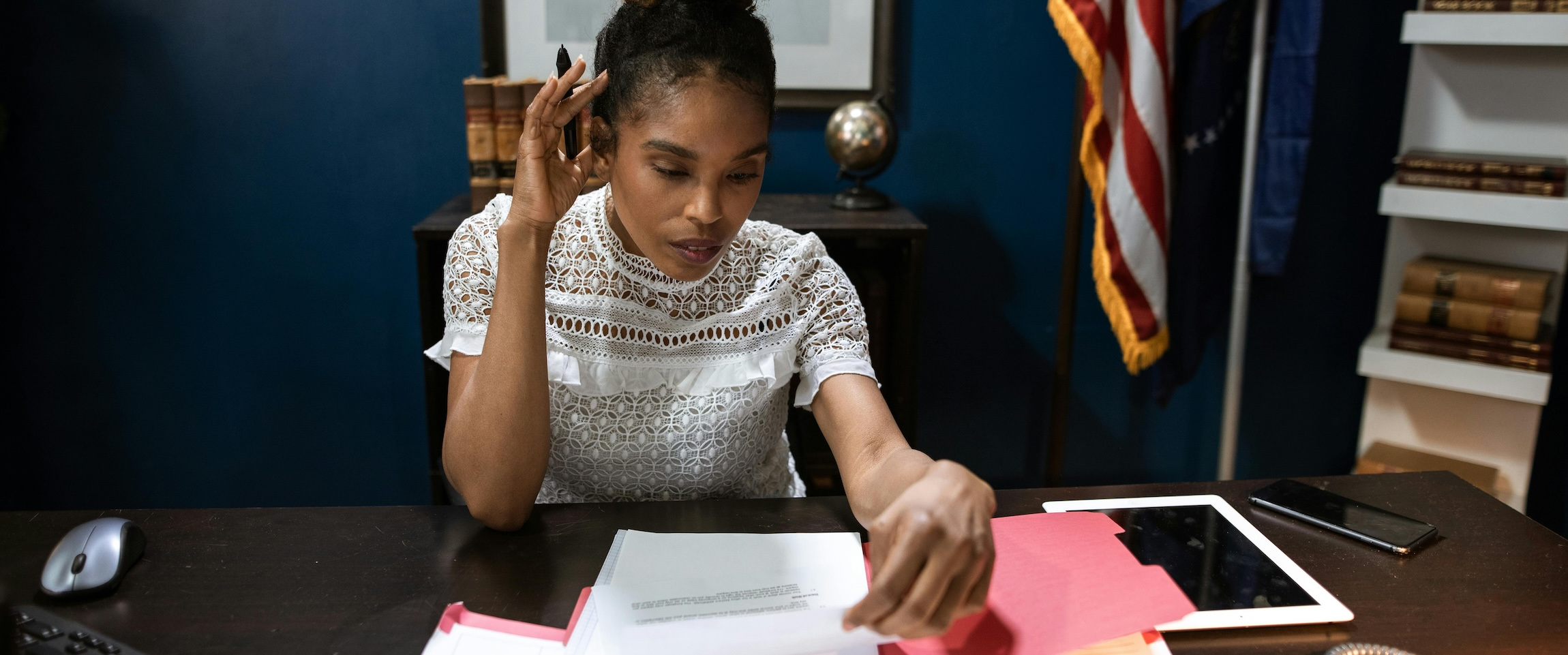 A woman sits in a government office with an American flag behind her, reviewing paperwork in a pink folder on her desk.