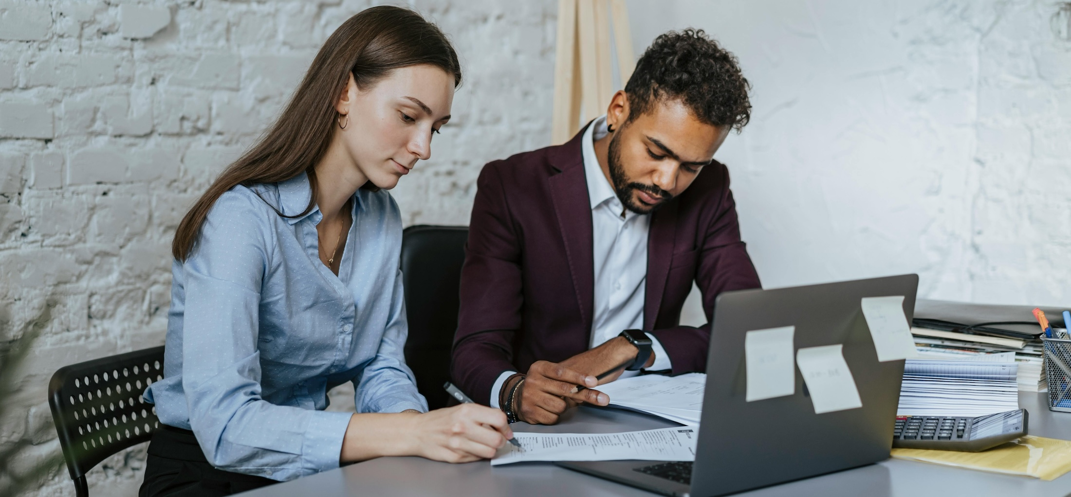 A man and a woman sit together at a table with an open laptop reviewing paperwork.