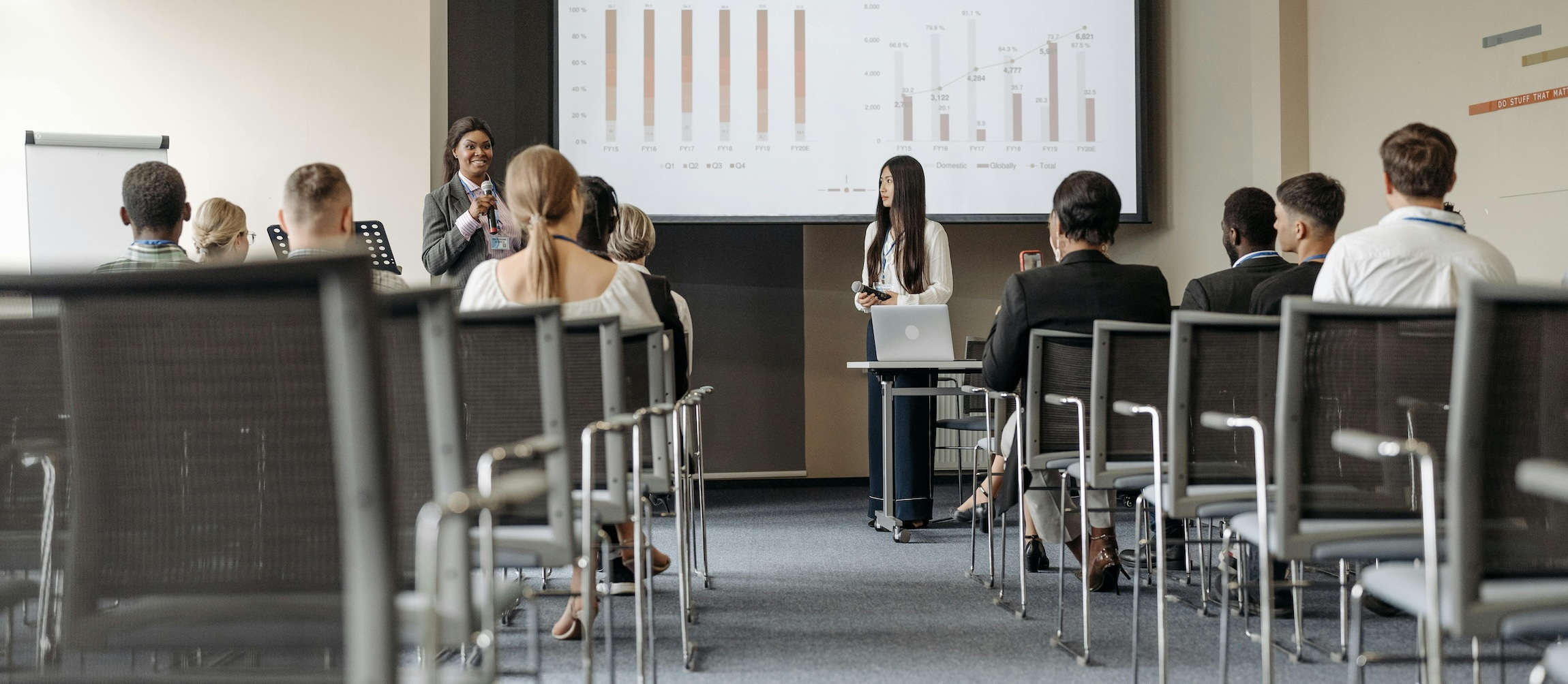 A woman speaks in front of a projected screen to a room of colleagues in business attire. Empty chairs are present in the back of the room.