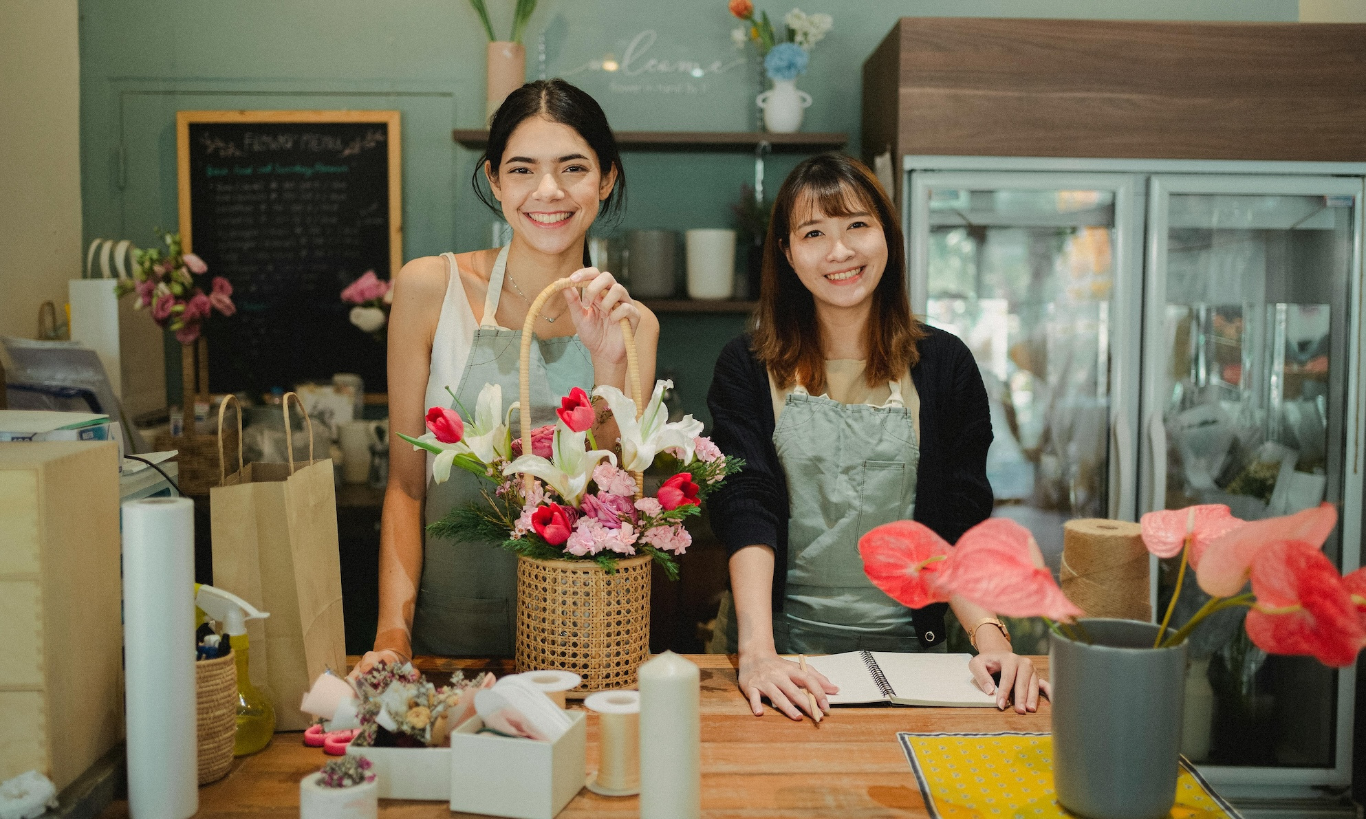 Two floral shop employees smile as one holds a floral arrangement and the other works on bookeeping.