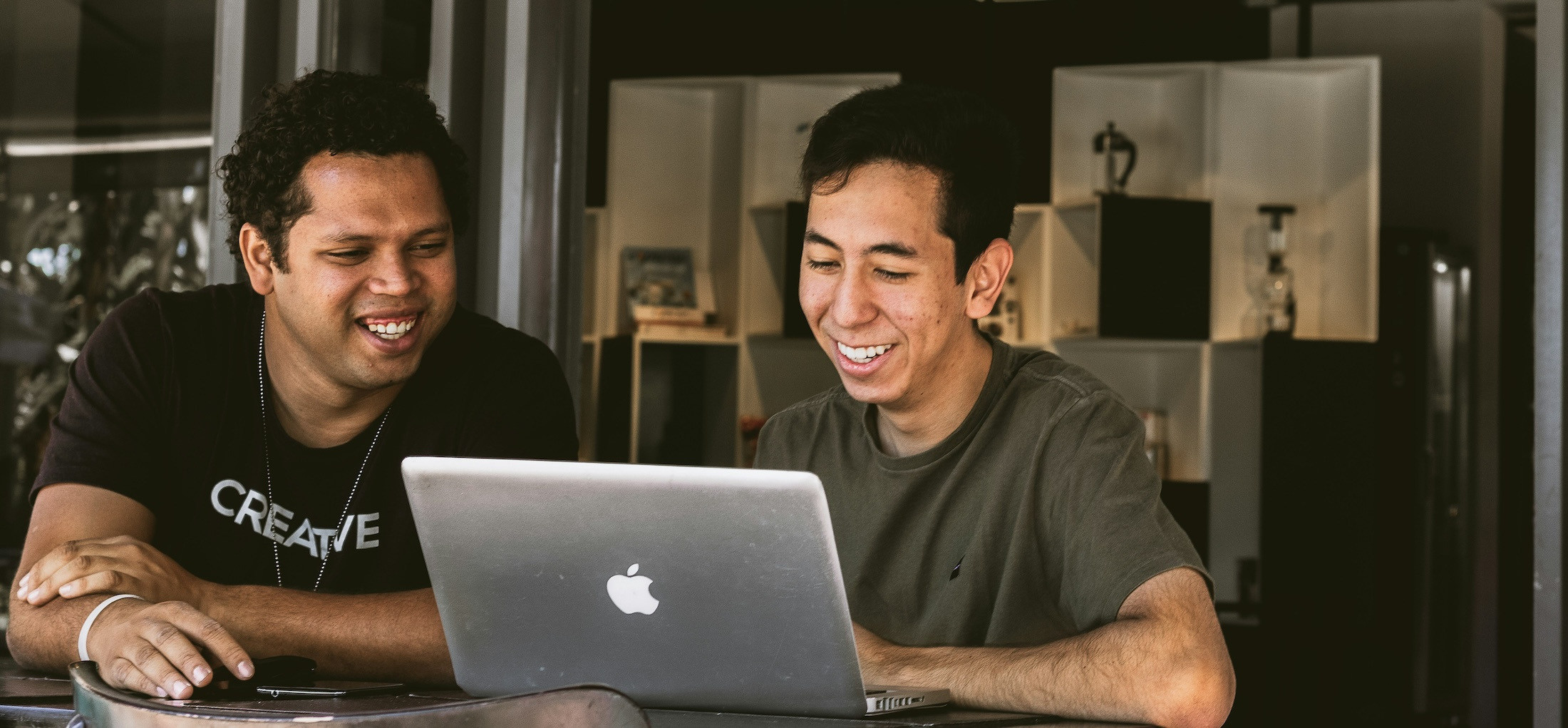 Two men wearing t-shirts look at a laptop screen and smile.