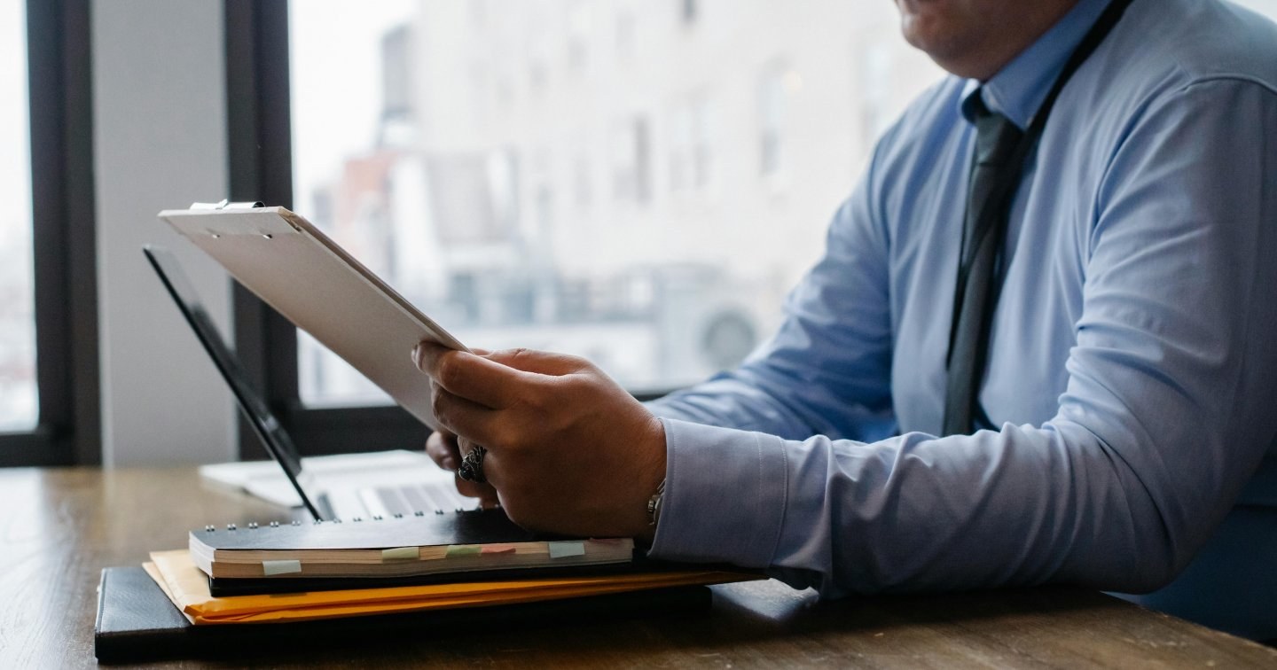 A man in a dress shirt and tie sits at his desk reviewing paperwork on a clipboard.