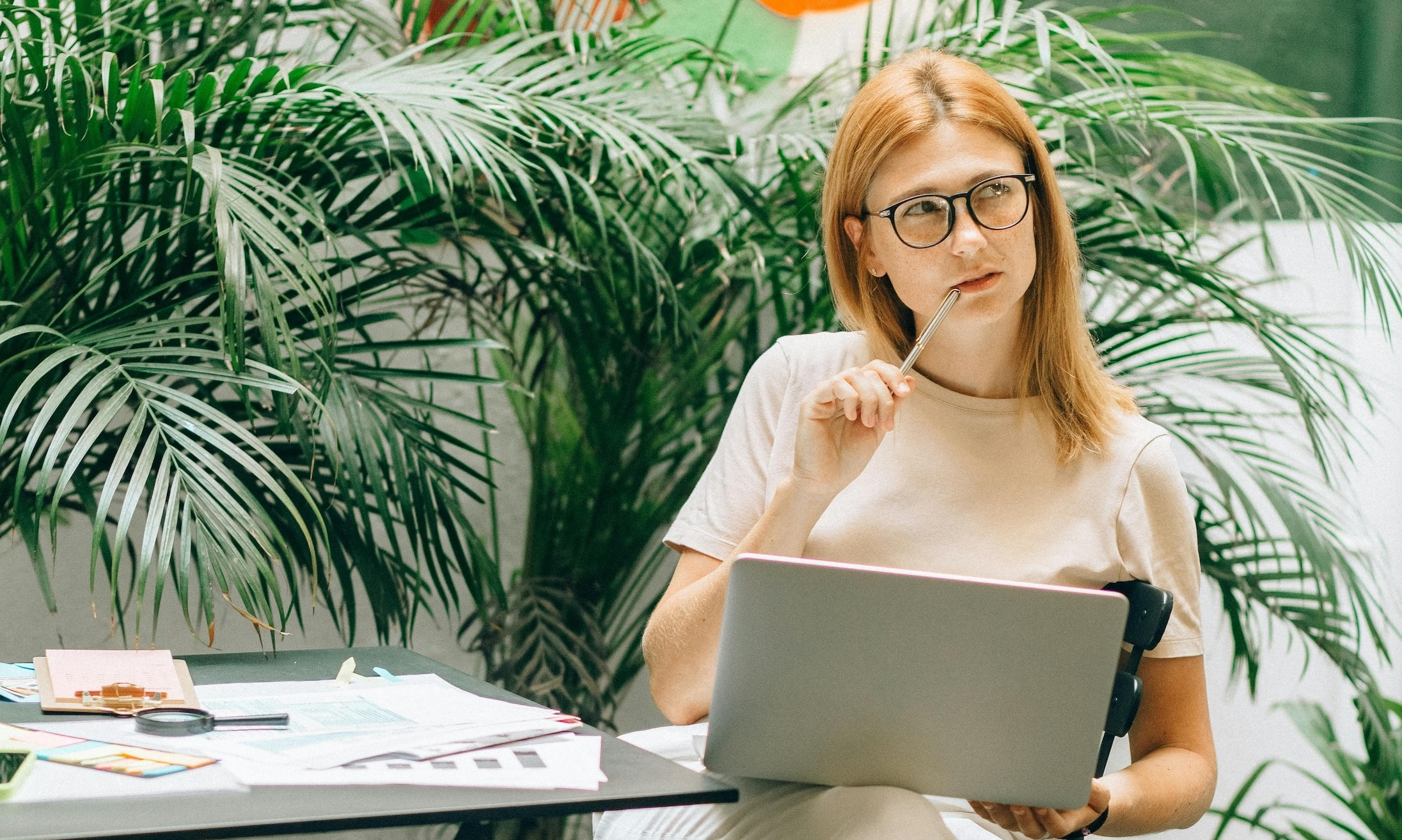 Woman wearing glasses sits in front of an indoor plant in a contemplative pose with a laptop in front of her.