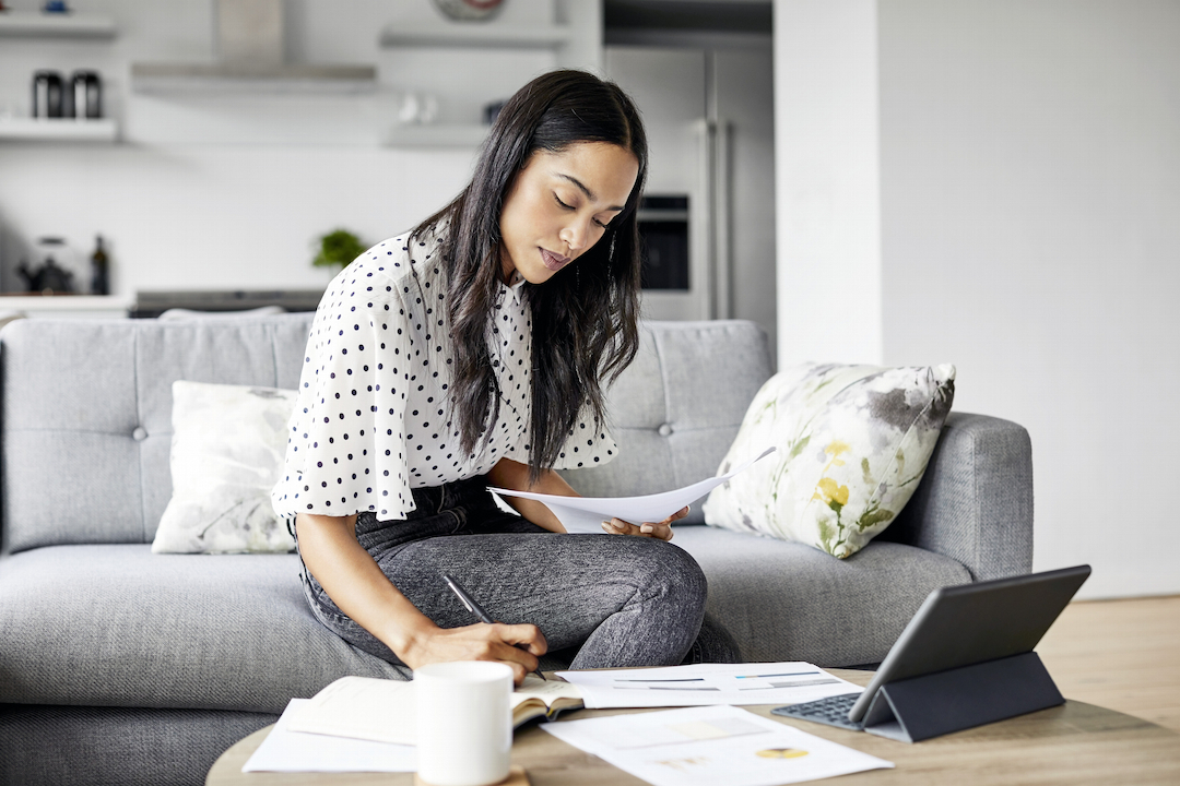 Woman sitting on a couch writing in a notepad next to other papers and a laptop.