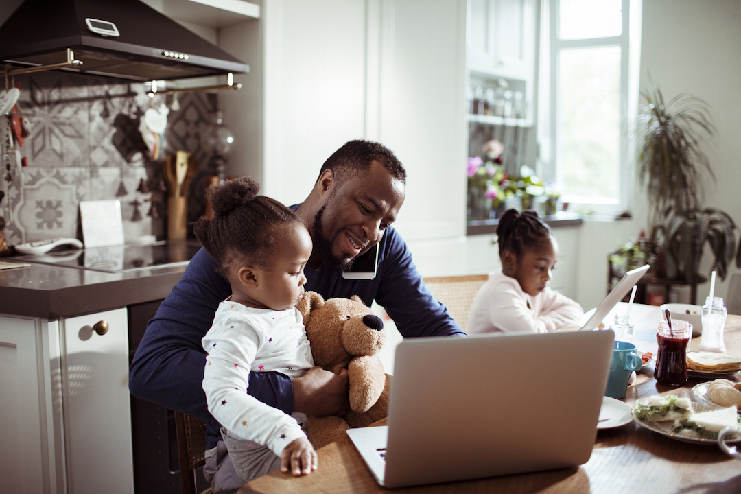 A man and two kids sitting at a table. The man is smiling at a laptop while talking on the phone. One kid is on his lap, the other is reading next to them.