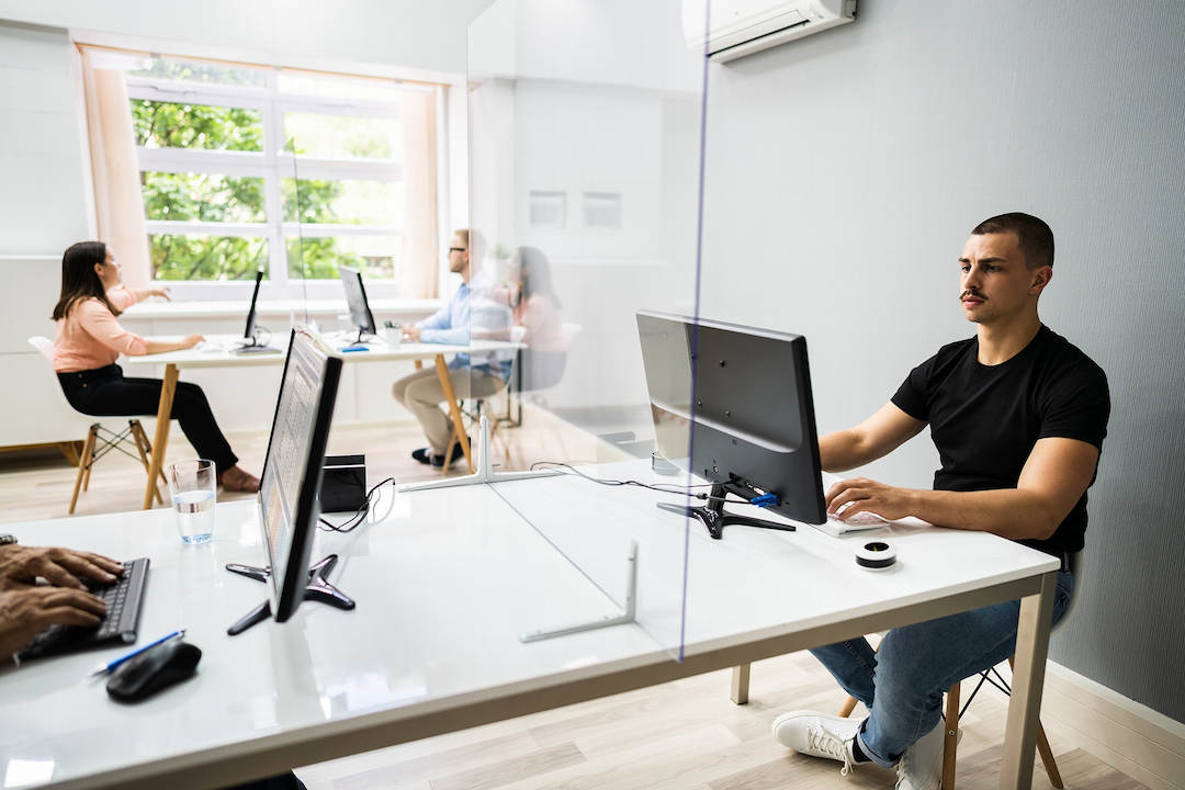 Two white desks with people sitting on the ends in front of monitors. There is glass separating the table in half, and 4 people working and talking.