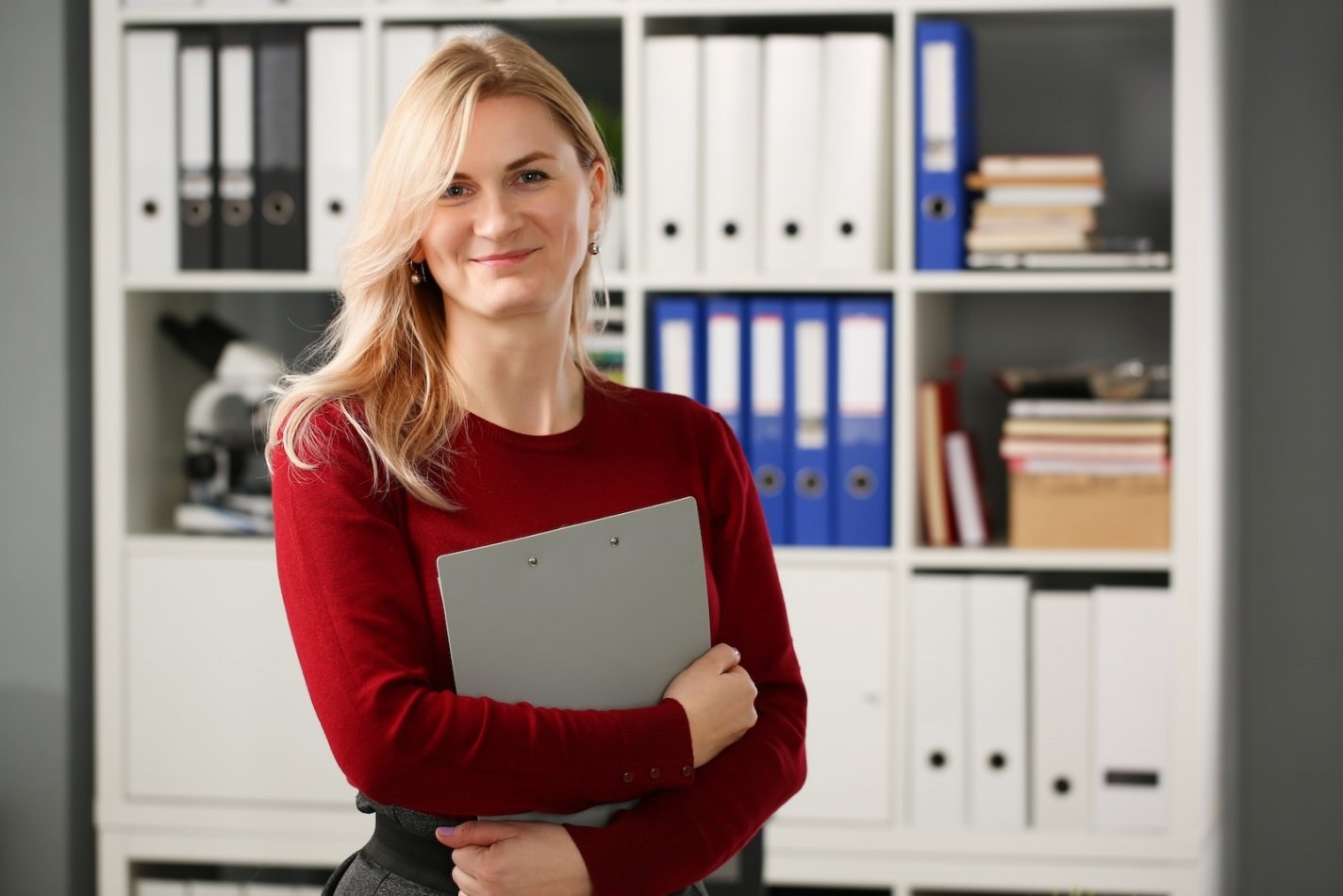 A smiling woman stands holding a clipboard in front of shelves filled with administrative binders