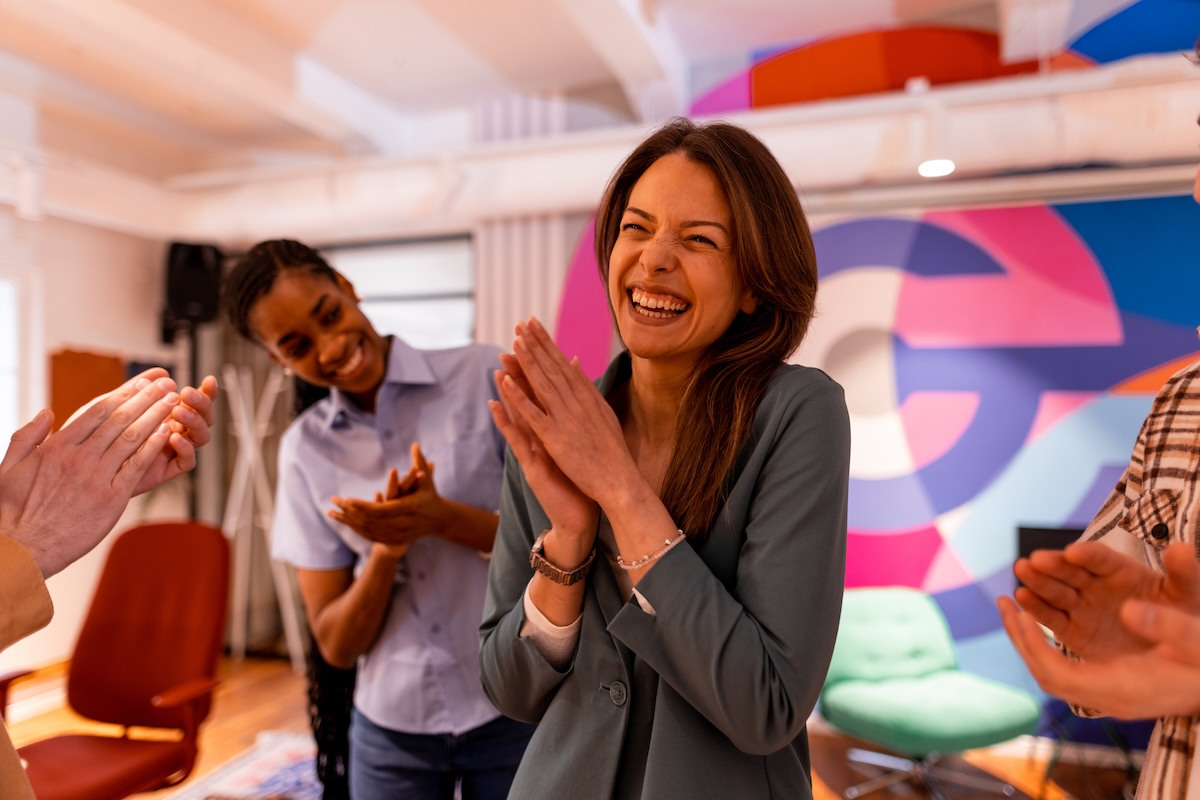 Woman smiles as her coworkers clap for her and recognize her at the workplace.