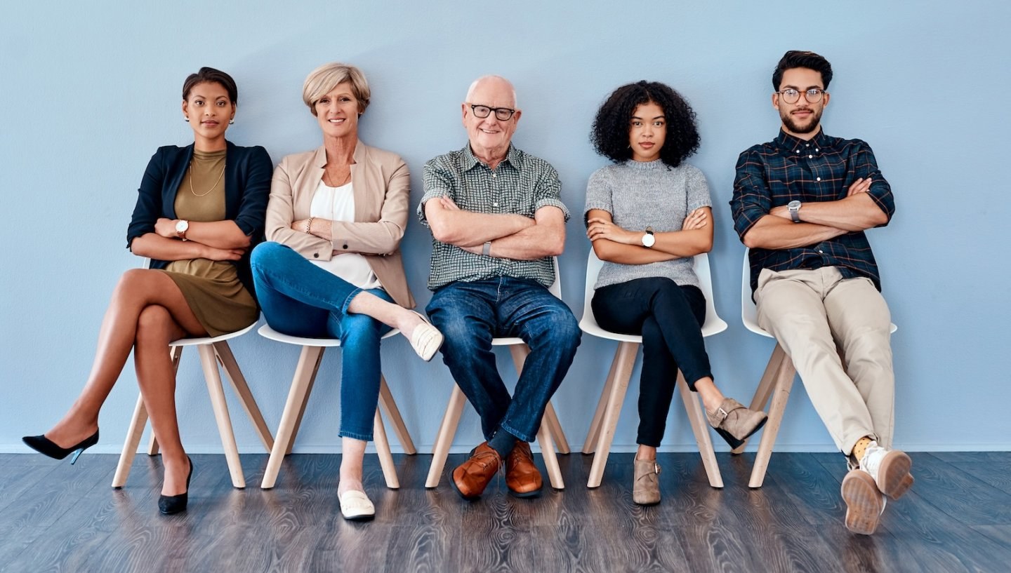 People of different age groups sit in chairs against a blue wall waiting to be called into an interview