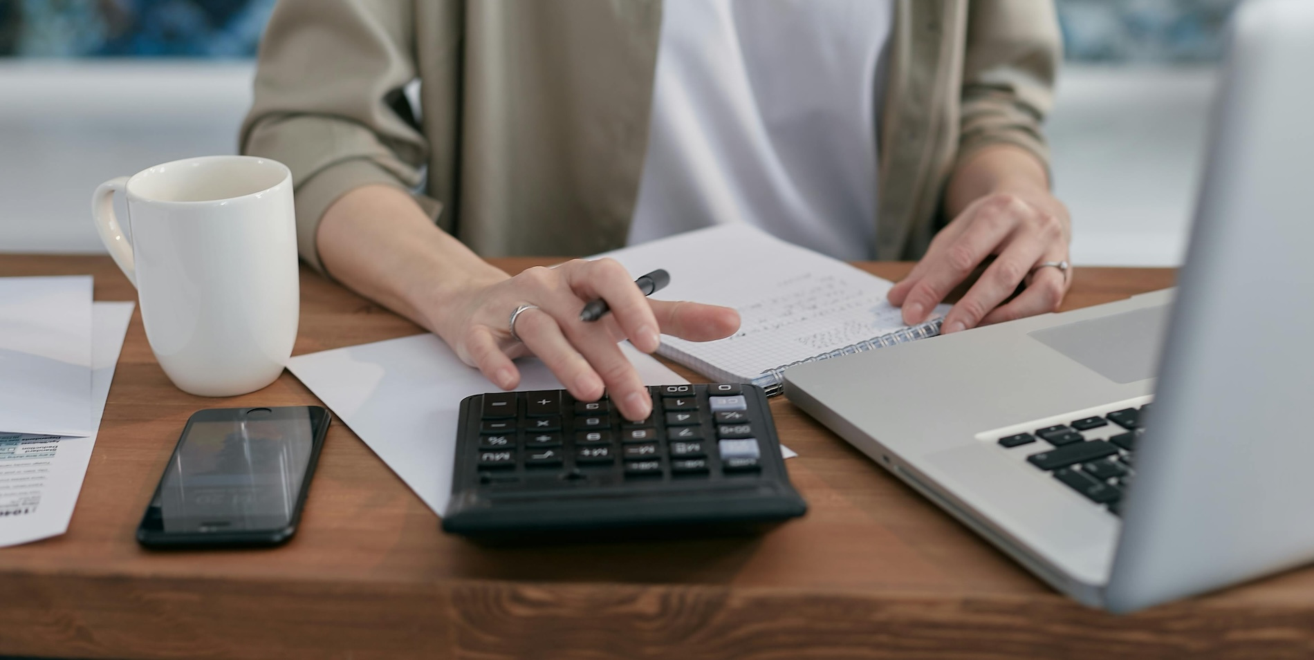 Person uses a calculator on a desk with an open laptop, mobile phone, and mug of coffee.