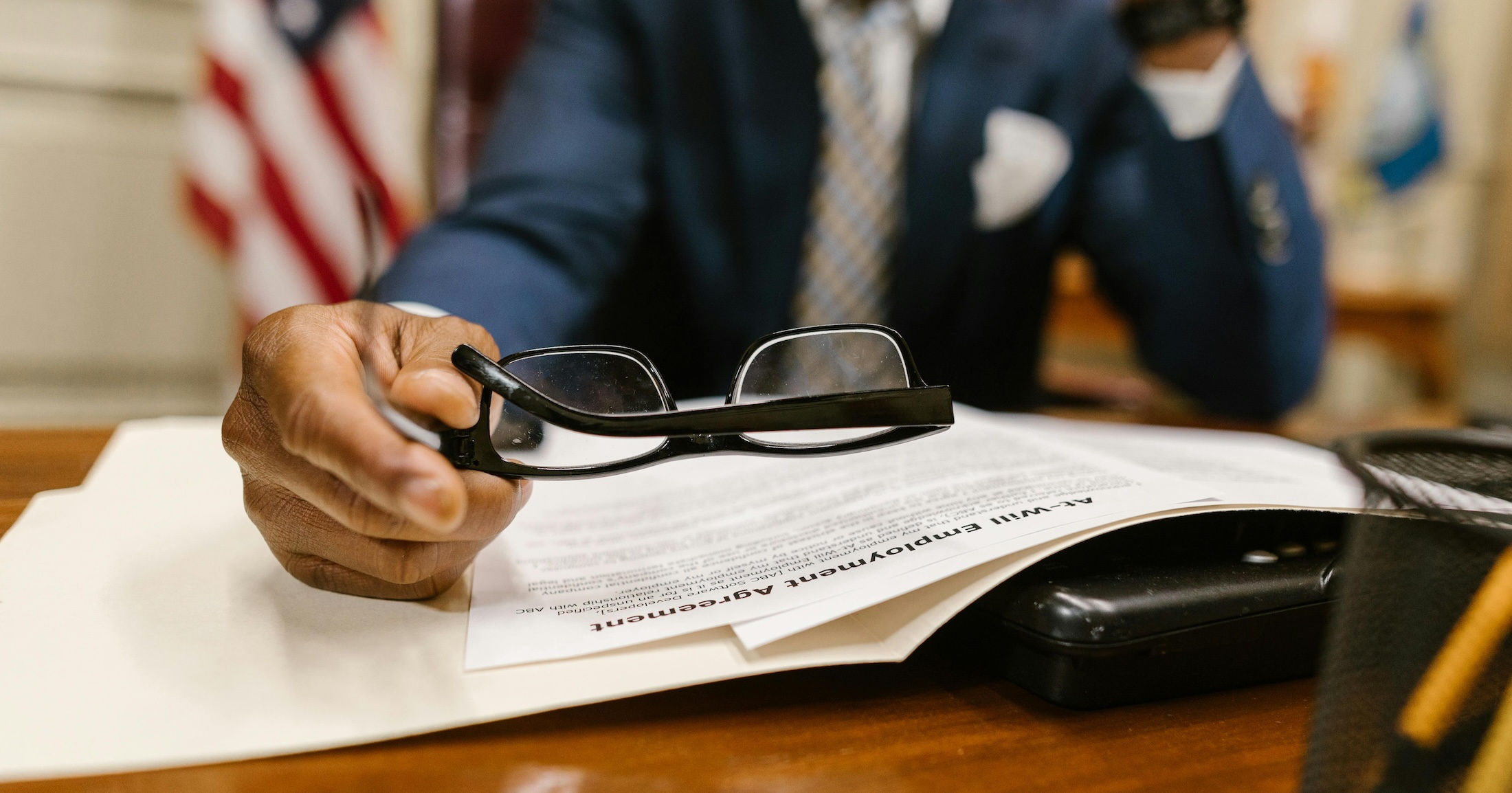 A man lays a pair of black framed glasses down on a pile of employment papers inside of a government office.