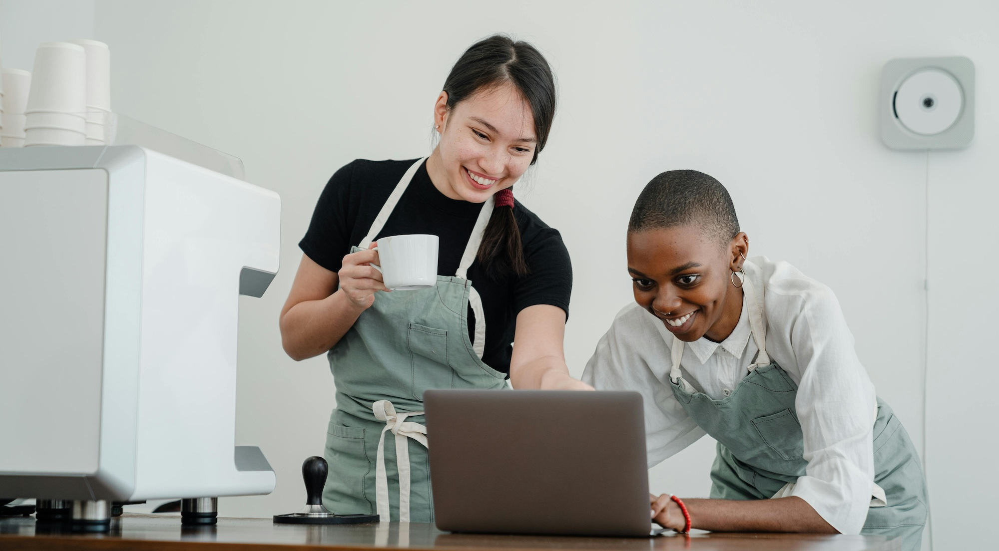 Two women wearing aprons, one holding a coffee cup, look at a laptop screen with smiles.