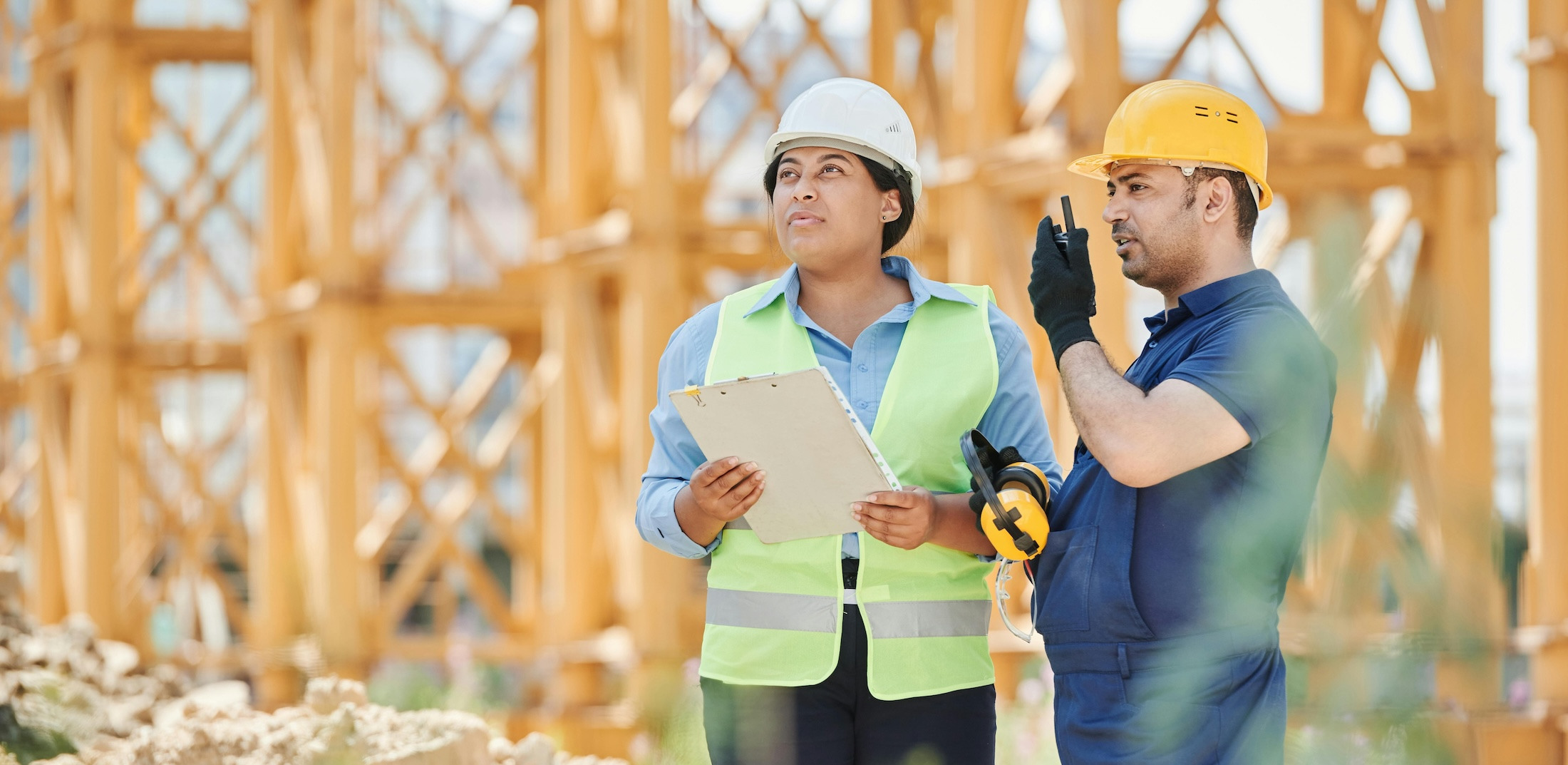 A man and a woman on a construction site wear helmets. The man talks into a walkie-talkie while the woman in a neon vest holds a clipboard.