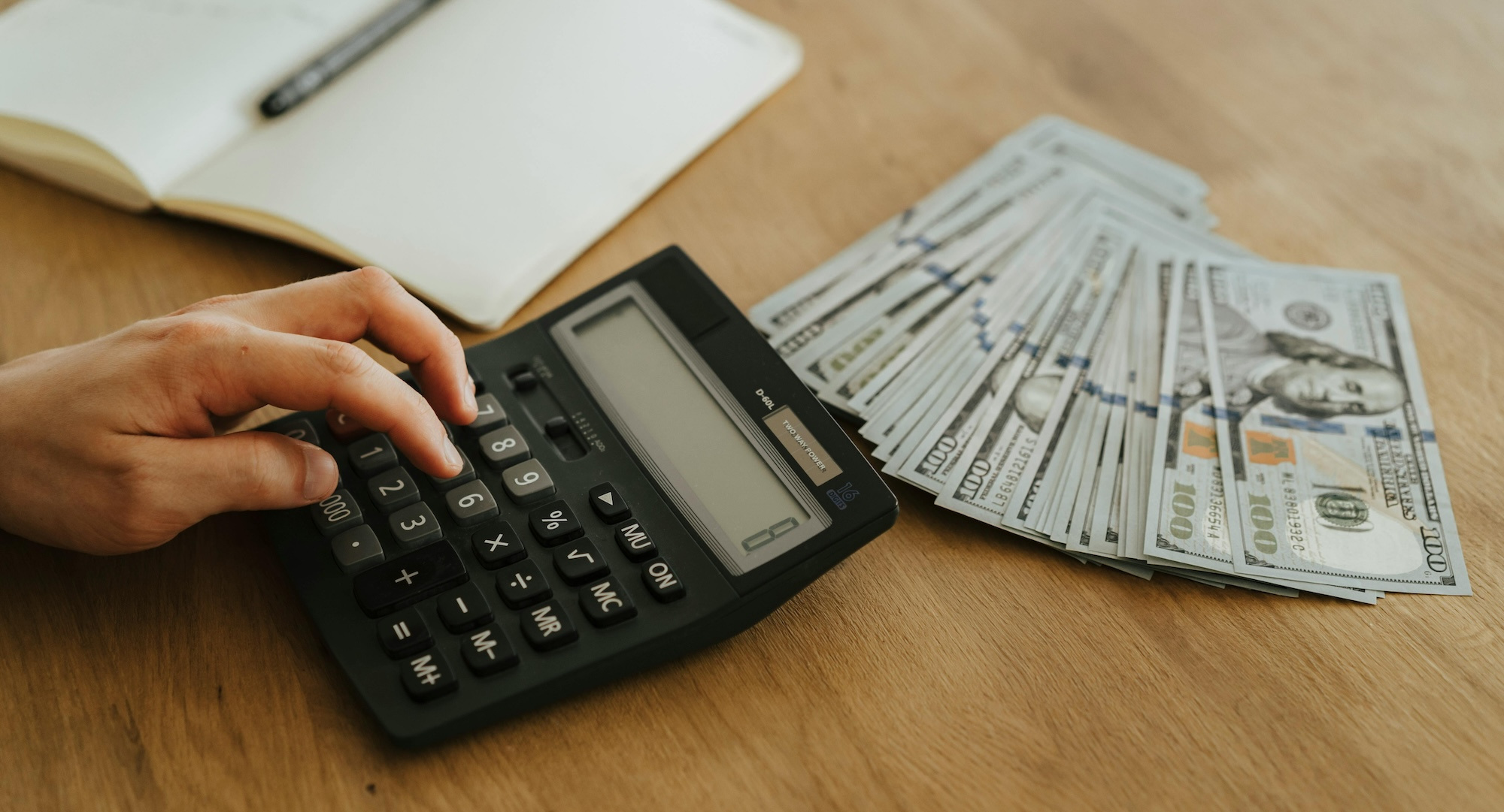 Fingers using a calculator on a table next to a pile of 100 dollar bills