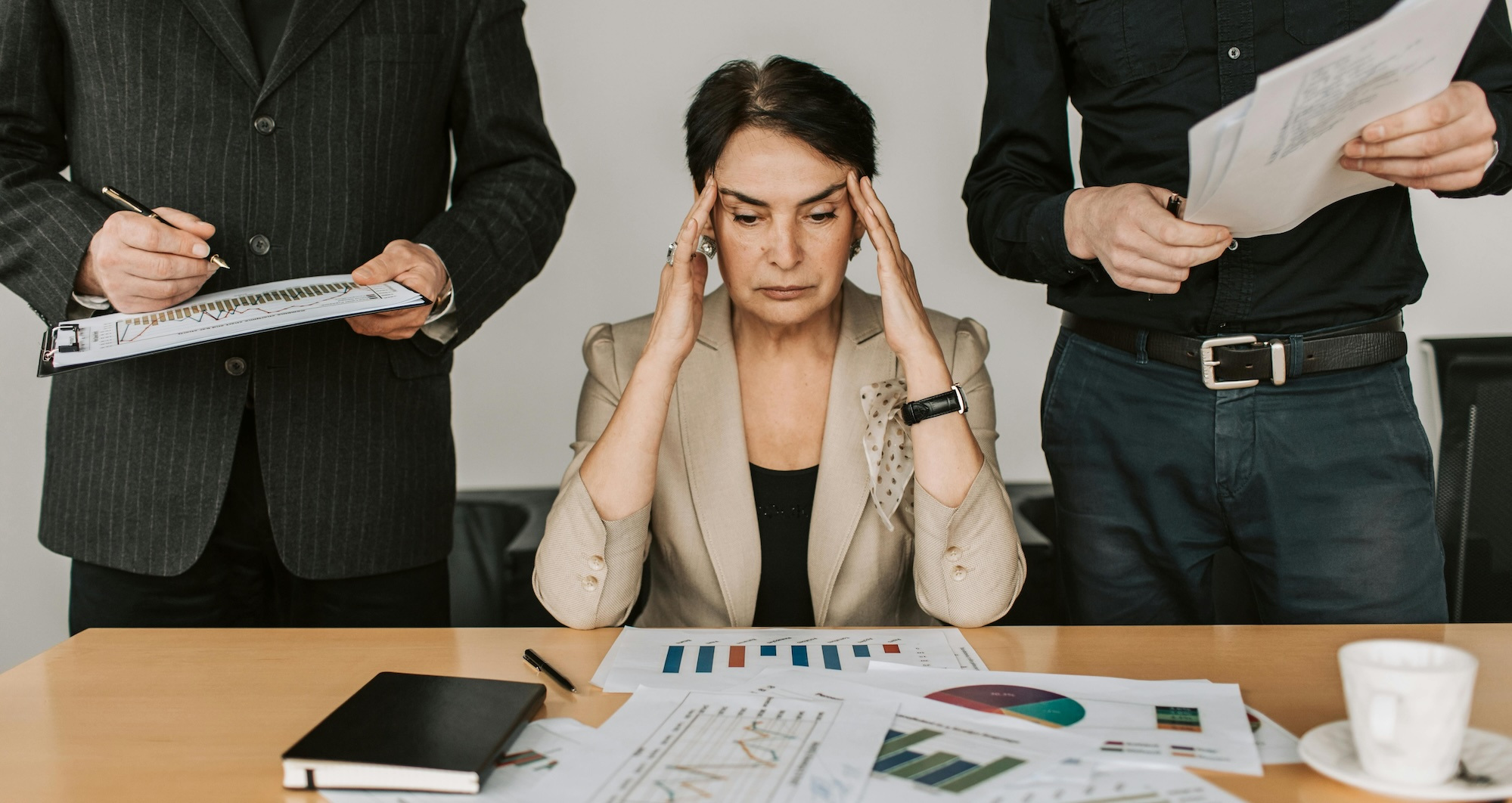 A woman in a beige blazer sits at a desk between two standing men, with her fingers on her temples looking overwhelmed in front of a table of scattered paperwork.