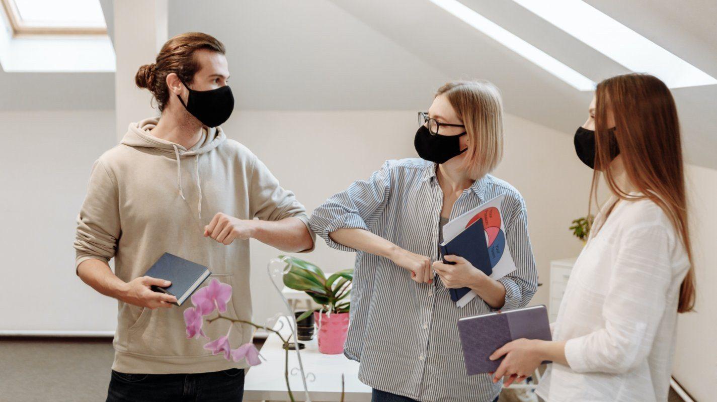 Three people wearing face masks, two of them are bumping elbows to greet one another.