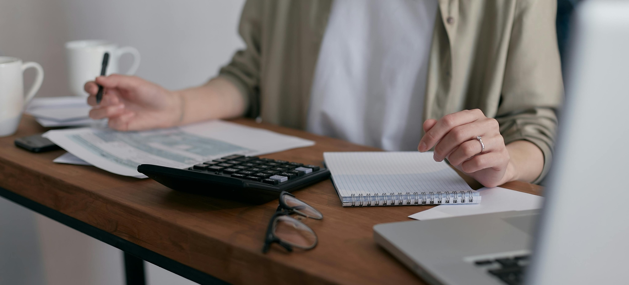 A person sits at a desk with an open laptop, notebook, calculator, and mugs while reviewing paperwork with a pen in hand.