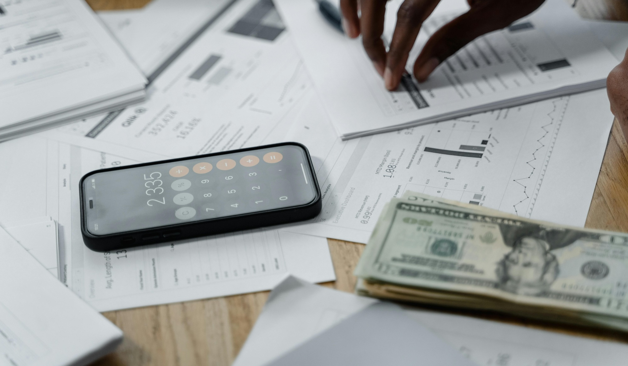 A desk covered in business paperwork, including an iPhone with the calculator app open and a stack of cash