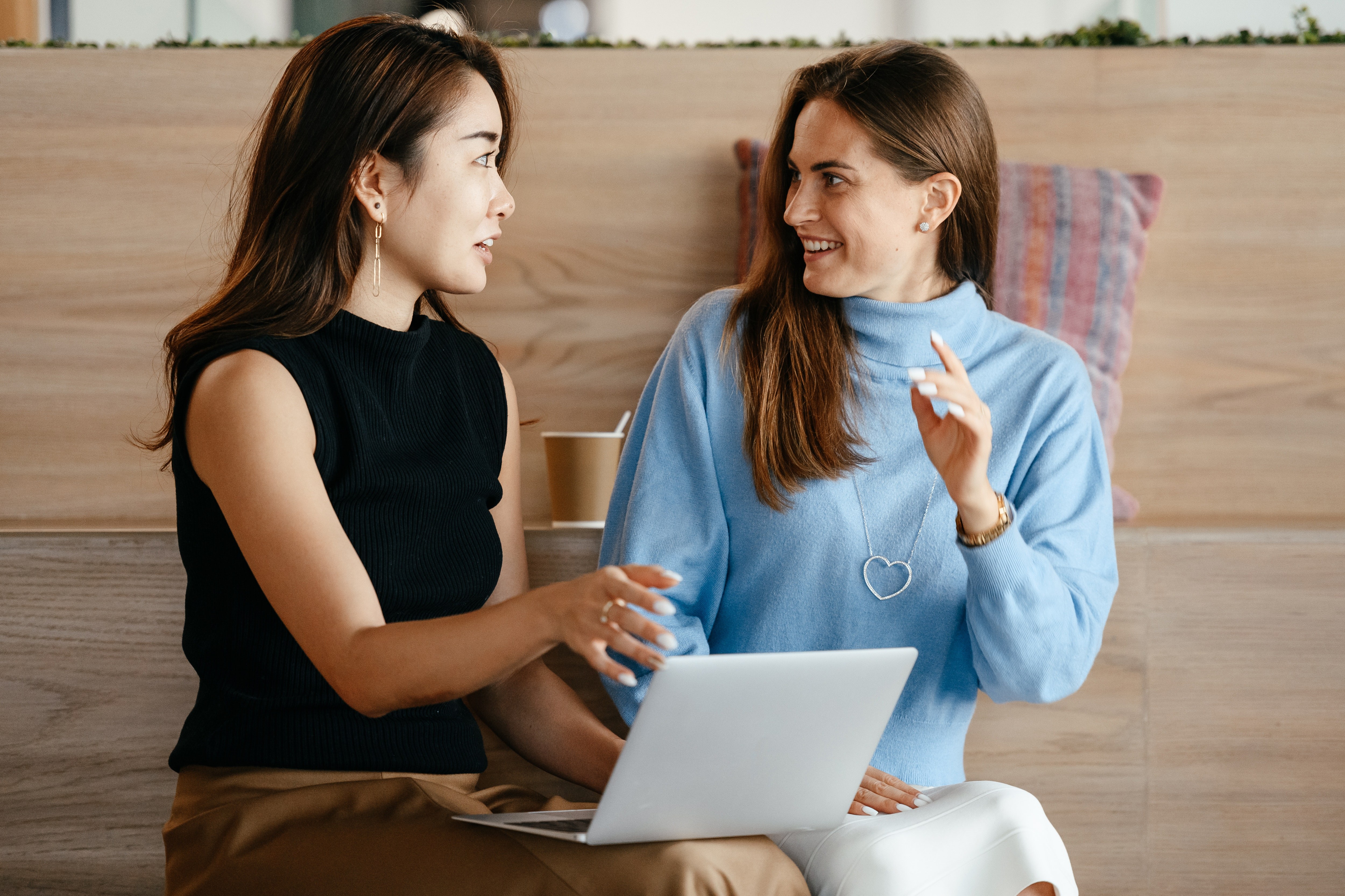 Two women talking and smiling at each other. One woman is wearing a black shirt and the other blue, and there is a laptop on the woman in black's lap.