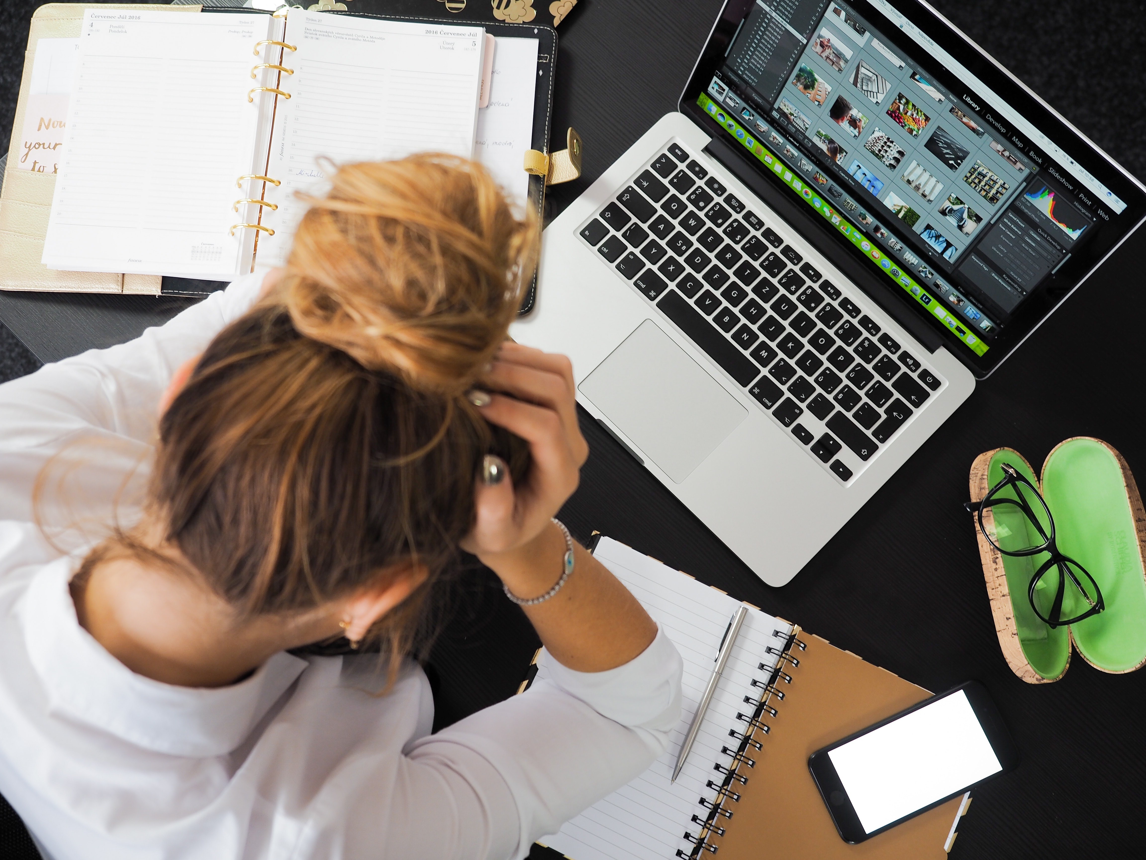 Woman with her head in her hands looking down at a desk with a computer, notepads, and glasses on the desk.