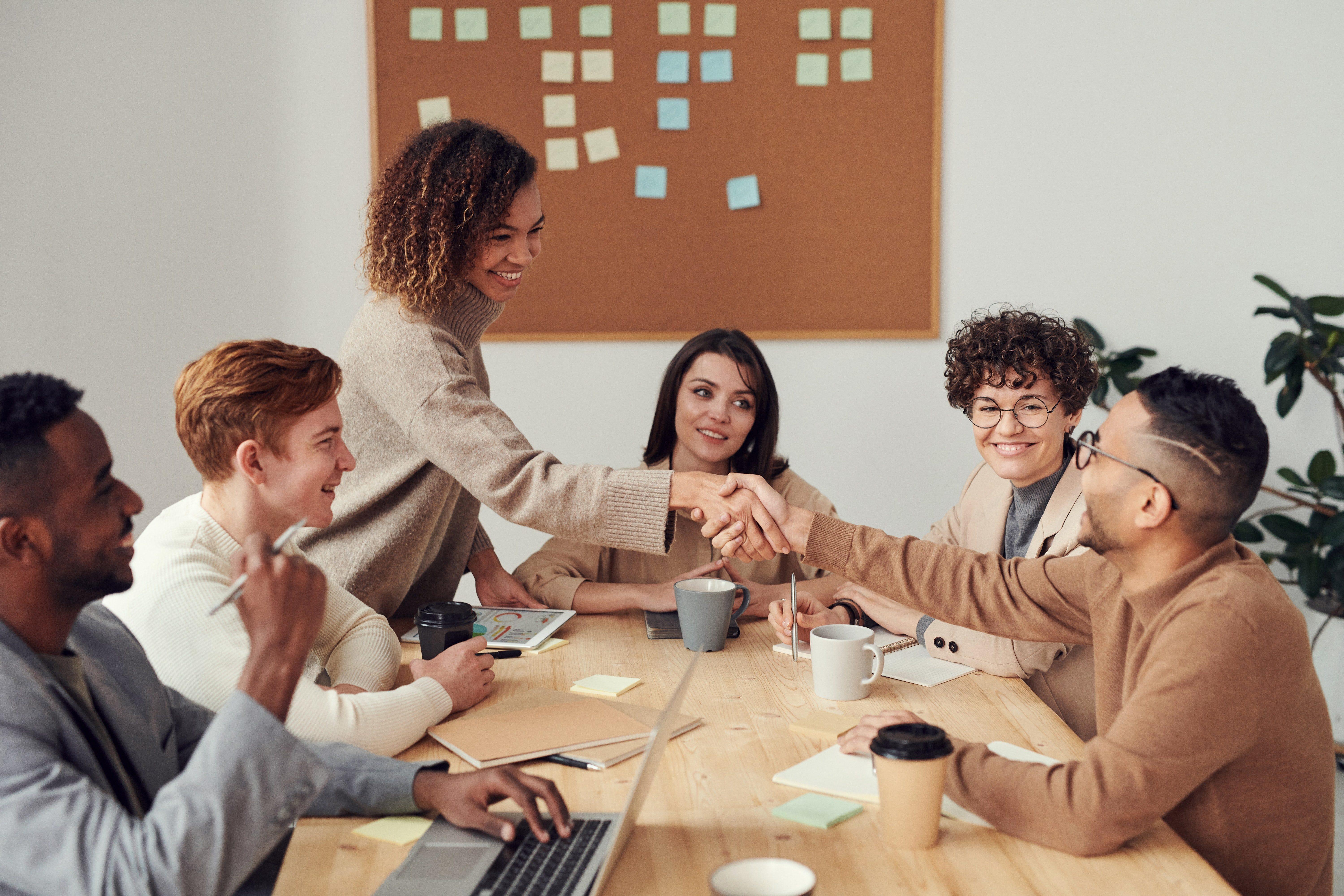 Group of people sitting at a desk, one woman is standing and shaking hands with a man sitting across the table.