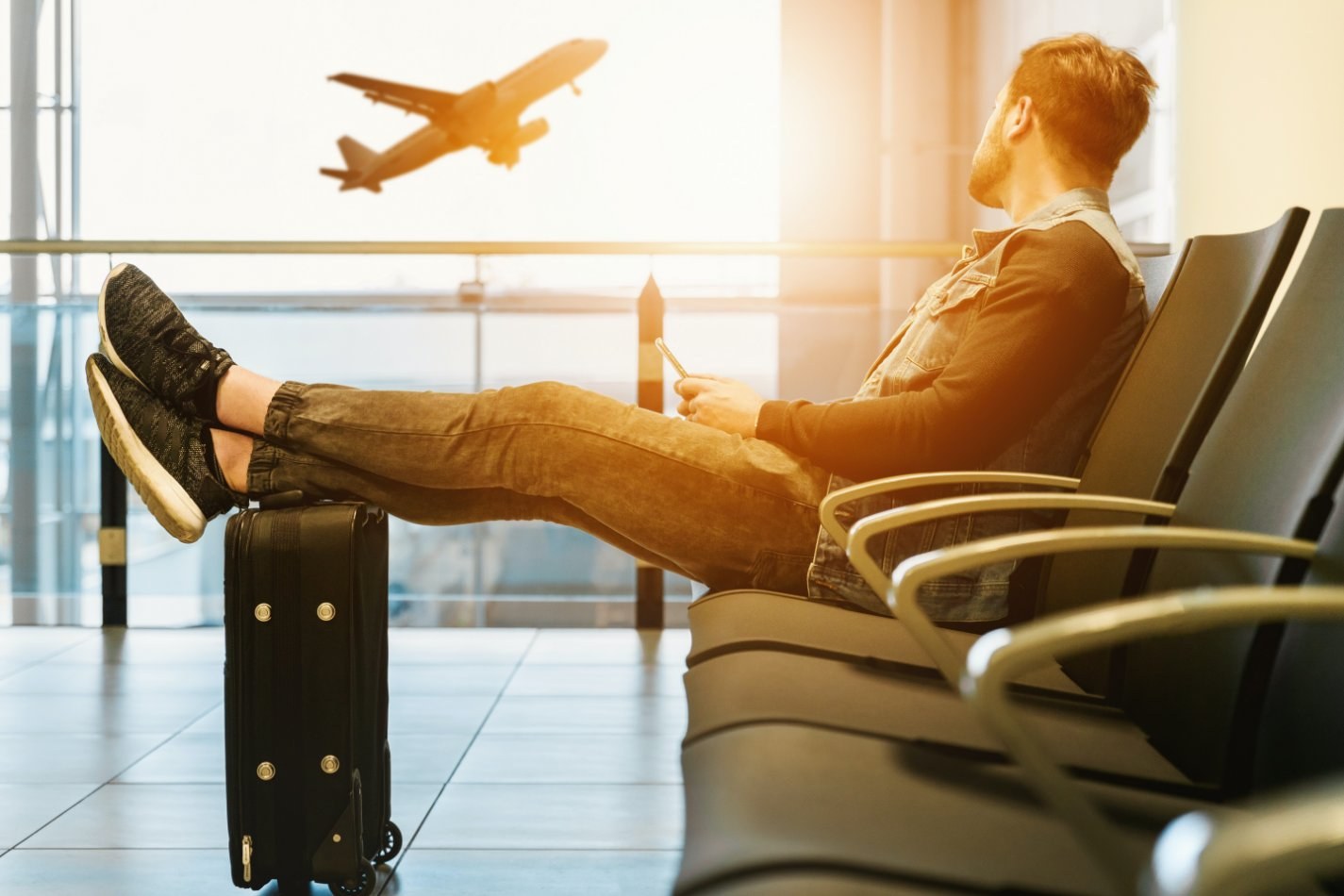 man at airport with feet on suitcase looking outside at plane taking off