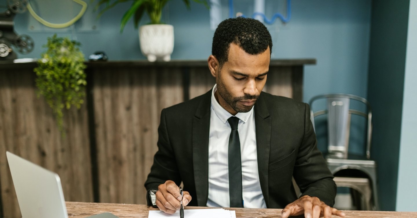 Man in suit sitting at desk, reviewing paperwork with pen in hand