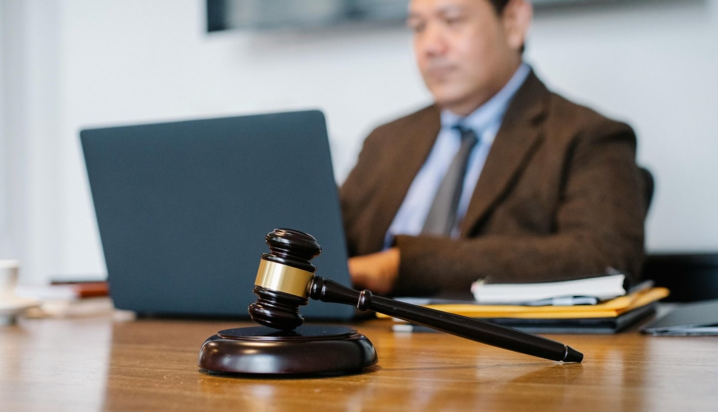 A man wearing business attire sits at a desk on his laptop while a gavel sits in the foreground.