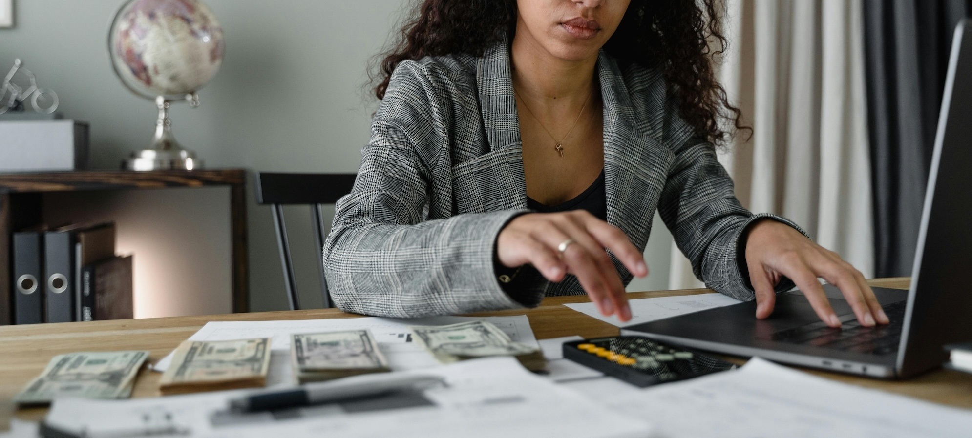 A woman wearing a blazer sits at a desk typing on a laptop with stacks of cash and a calculator on her desk.