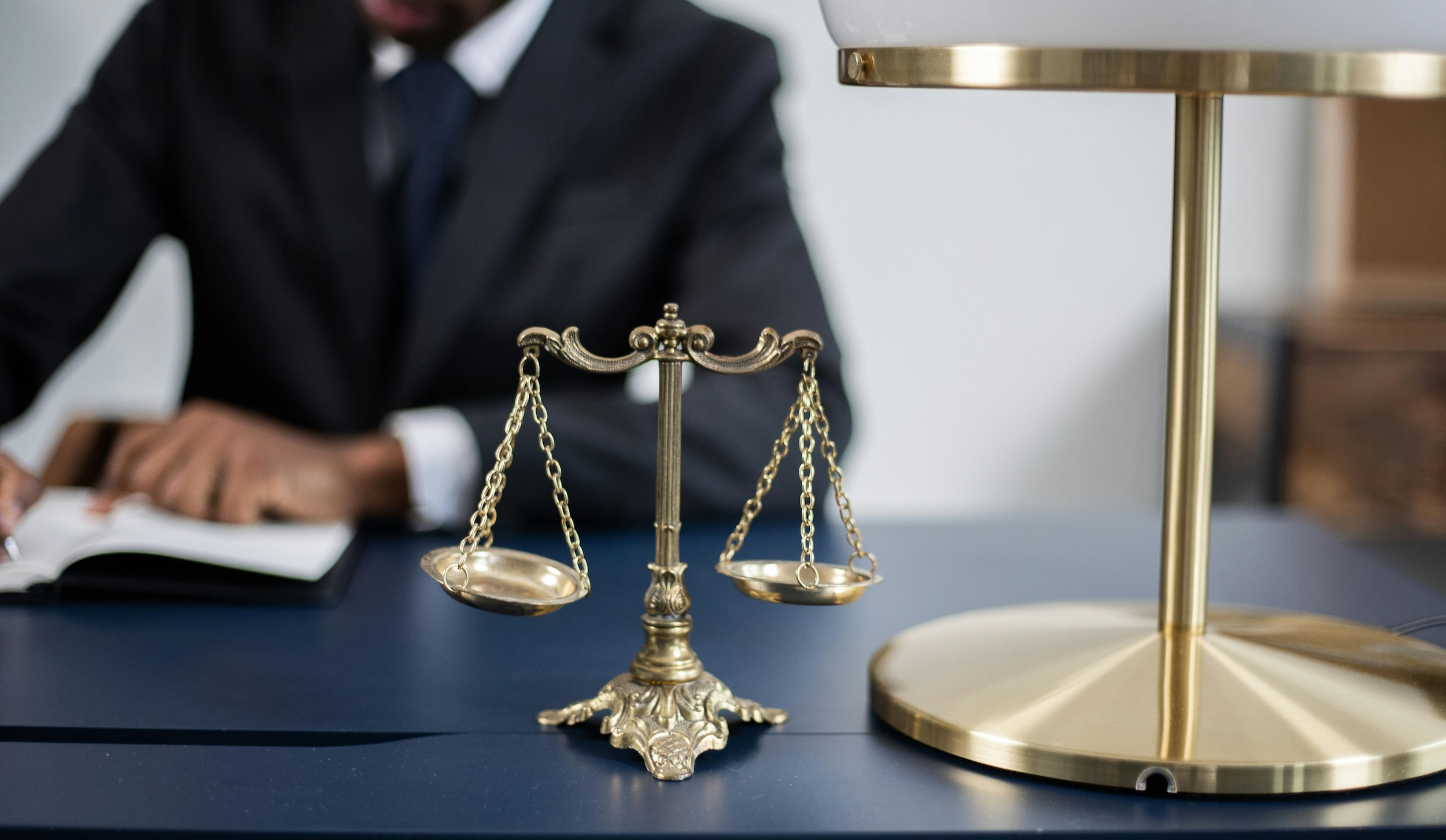 A man in a suit sits in the background with the image focused on gold scales of justice sitting next to a gold lamp on a desk.