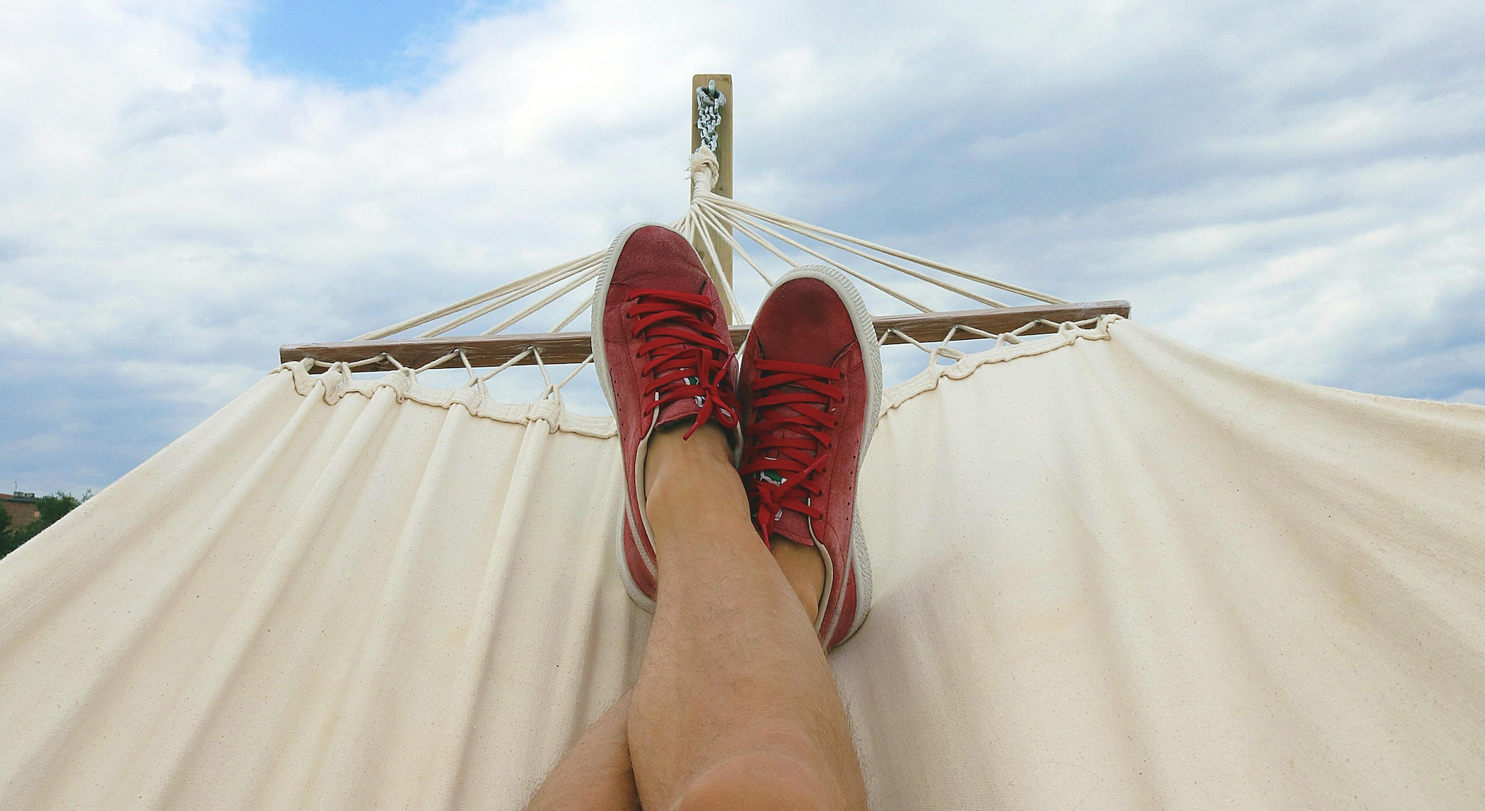 Person wearing red sneakers reclines their feet in a hammock with a cloudy blue sky in the background.