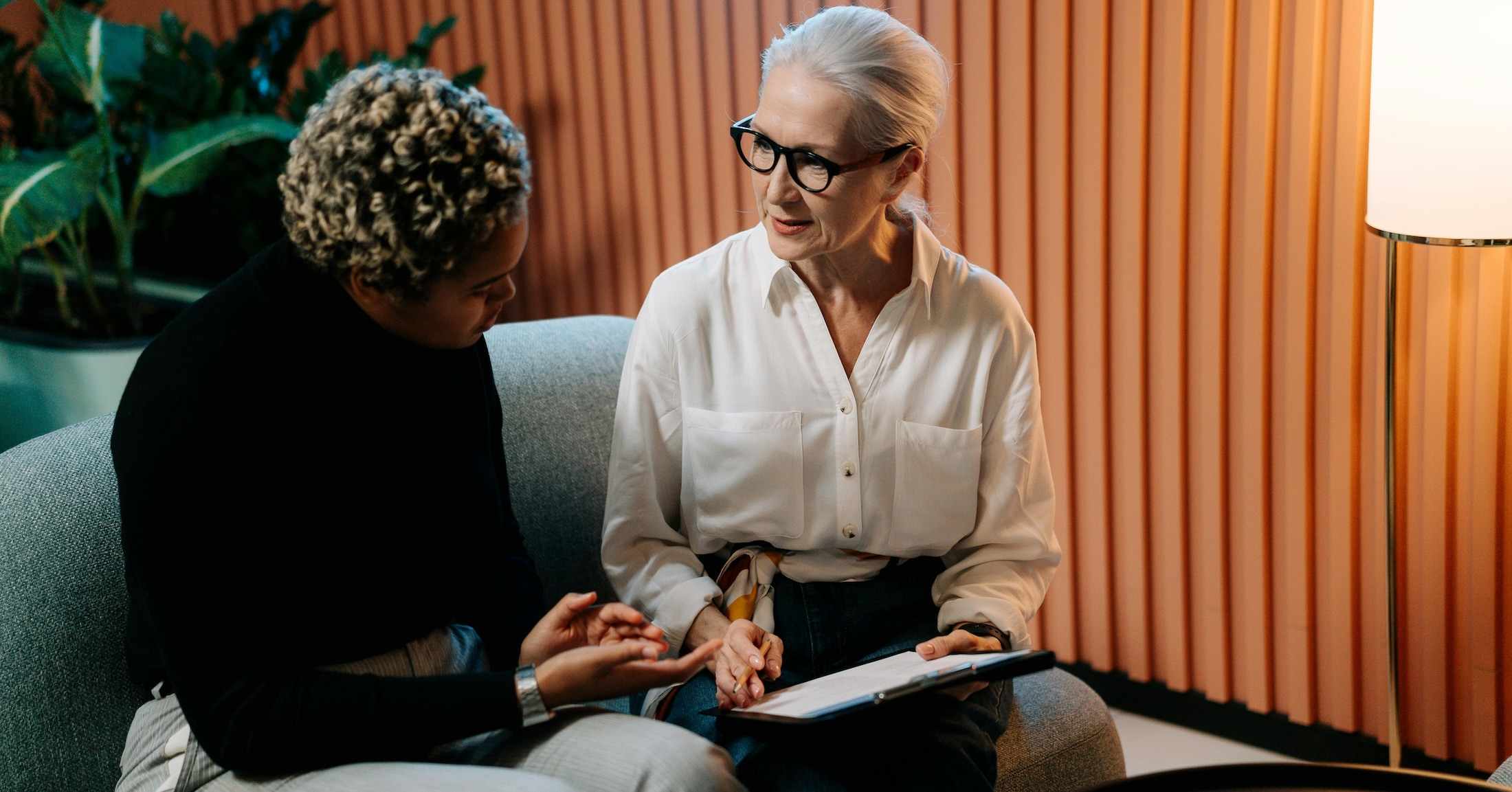 Two women sit on a couch at work looking at paperwork and discussing with each other.