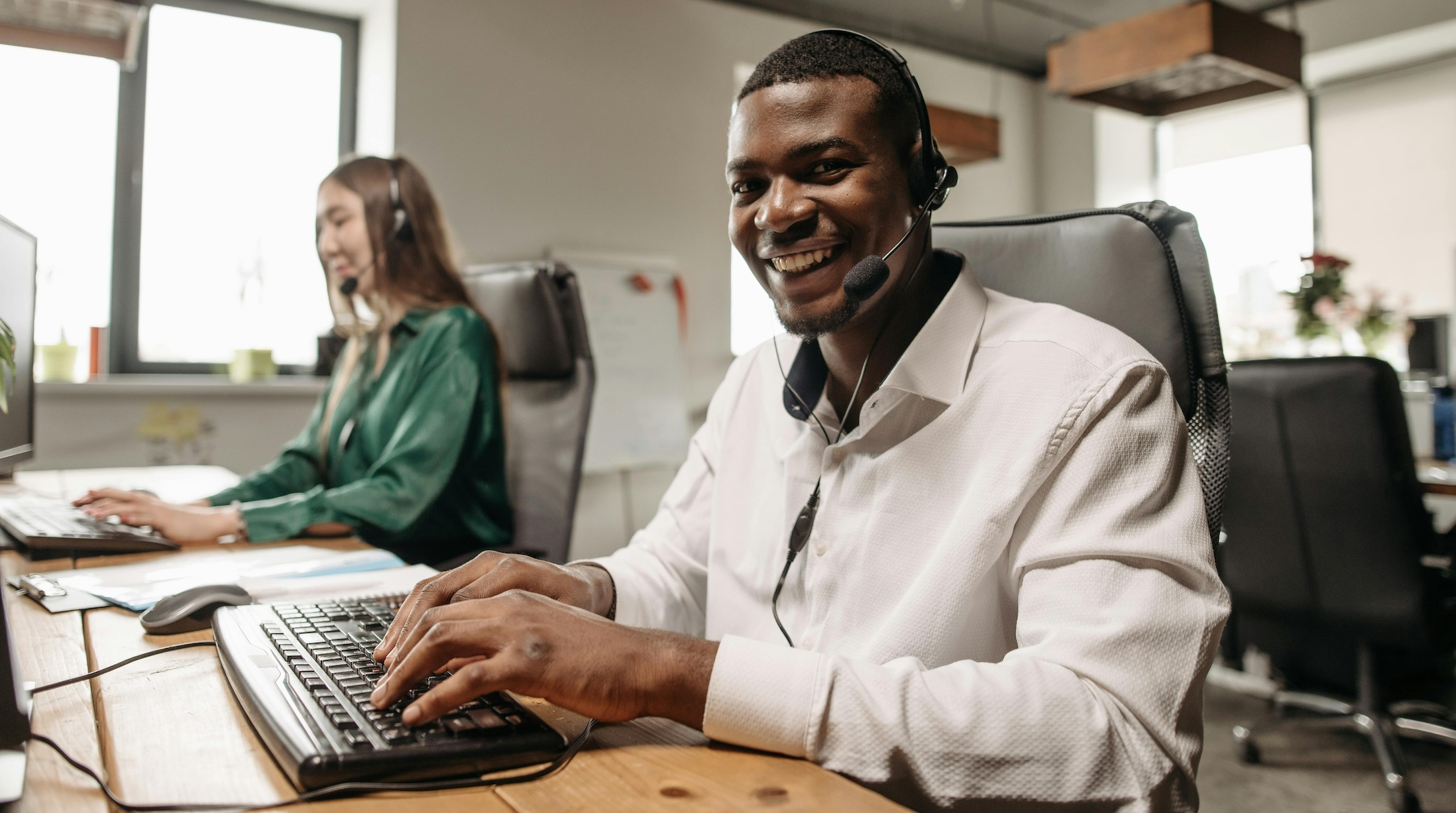 Man using a computer from a modern call center, wearing headset and smiling.