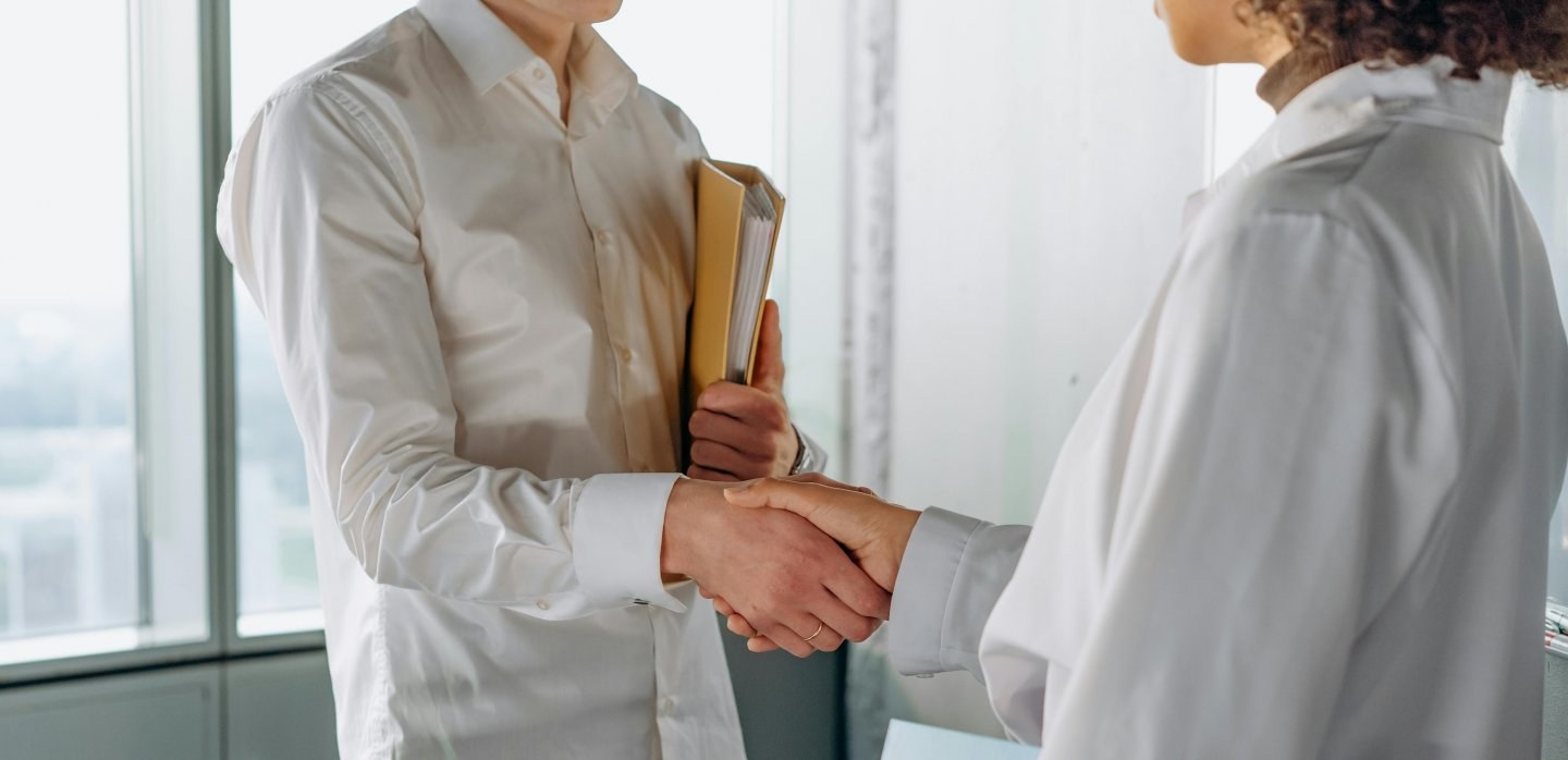 Two people in white shirts shaking hands in an office. One carries a yellow binder.